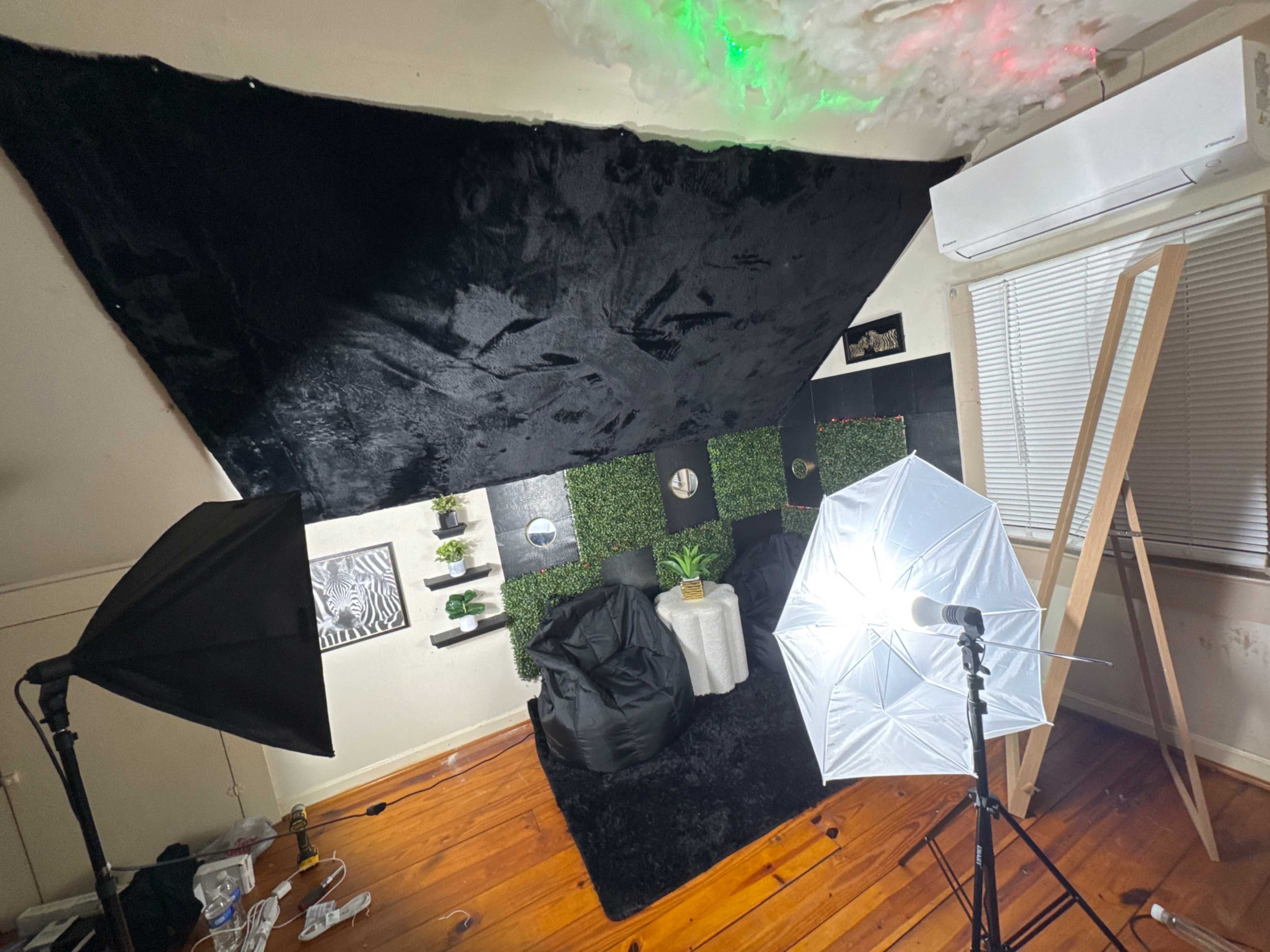 The image shows a small room setup for photography, featuring black and green decor, bean bags, light stands, and an air conditioning unit.