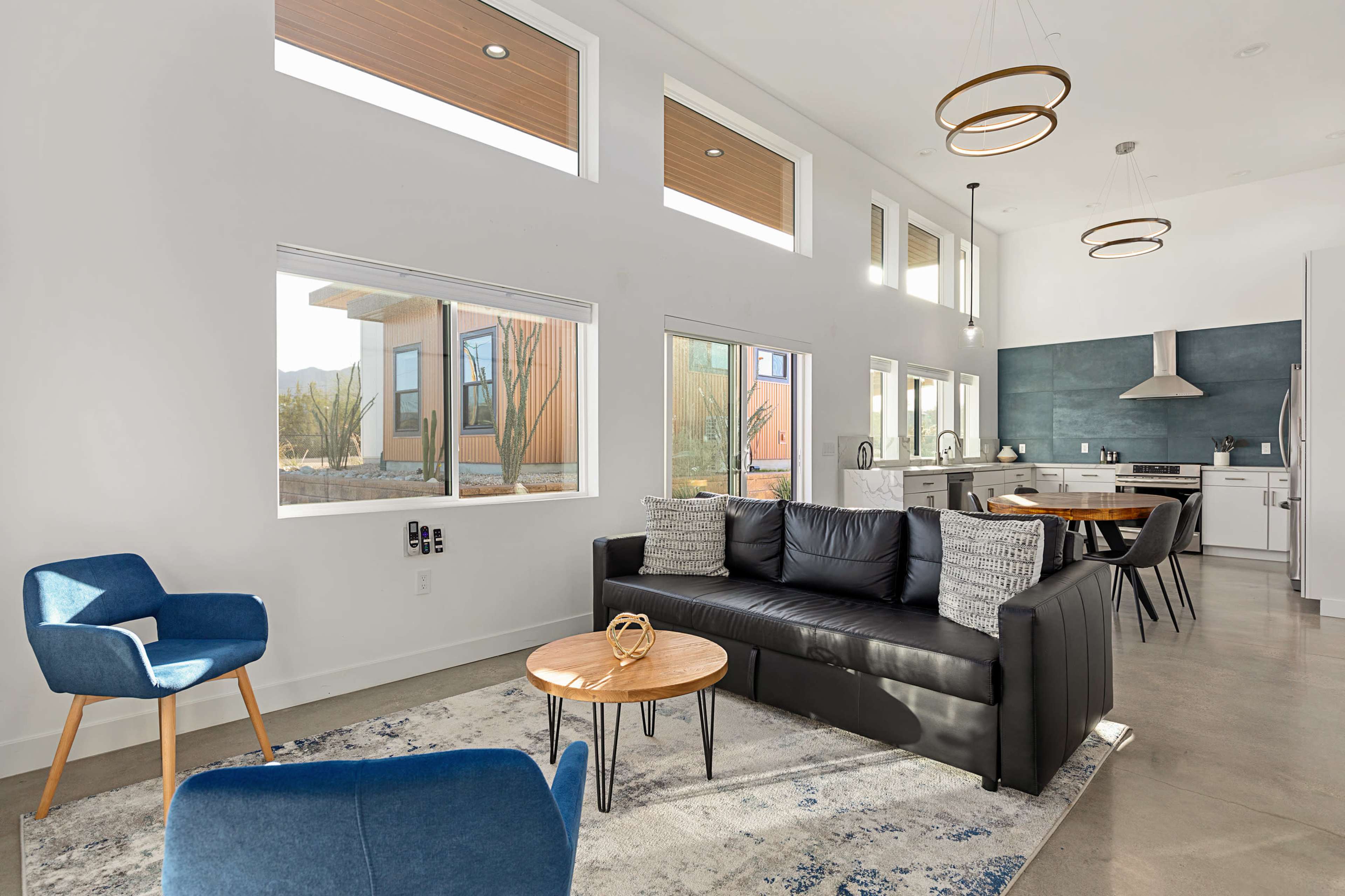 The image shows a modern living room with large windows, minimalist furniture, and a kitchen area visible in the background.