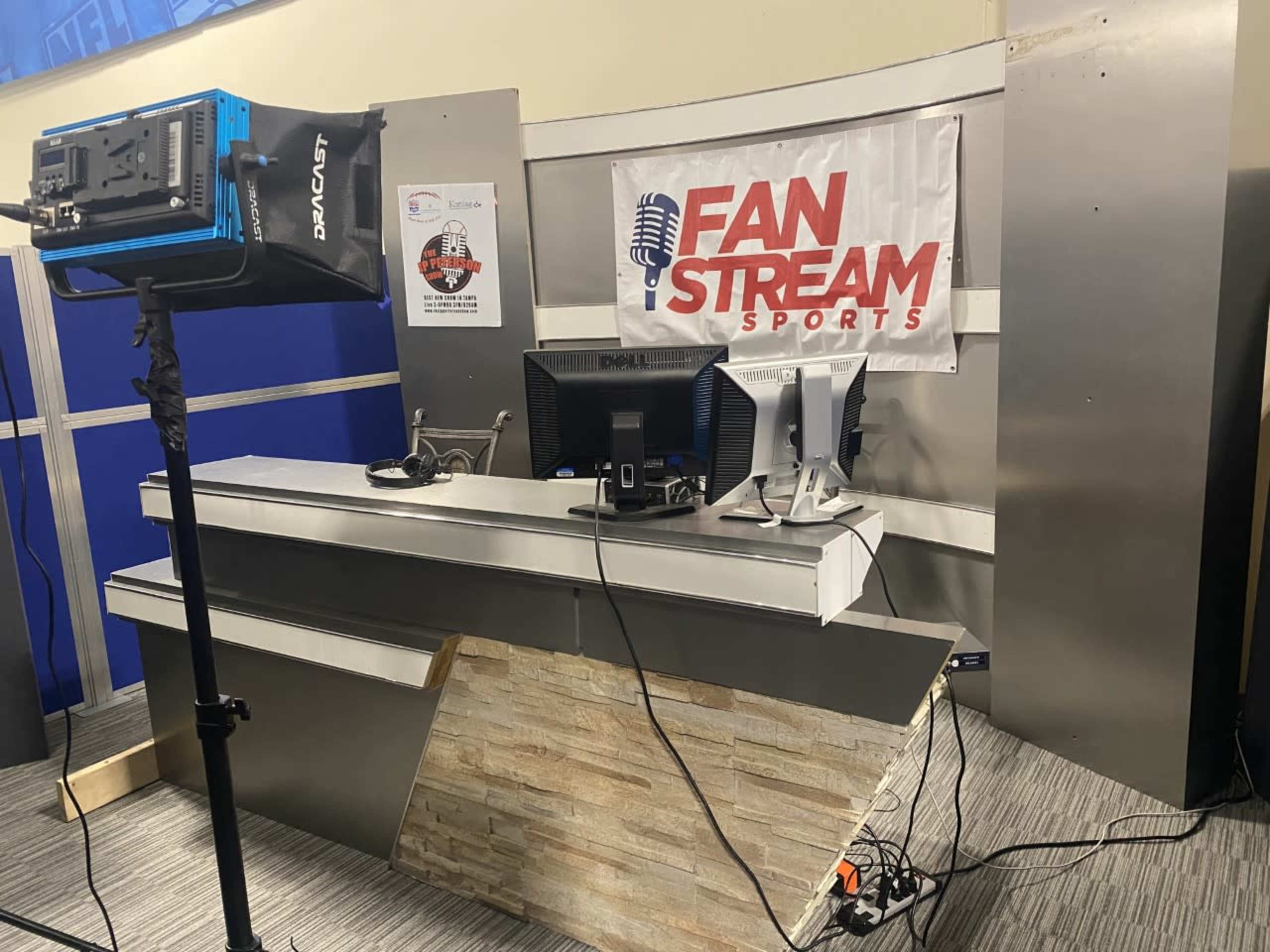 A sports broadcasting studio setup featuring a desk with monitors, a microphone banner, and a film light.