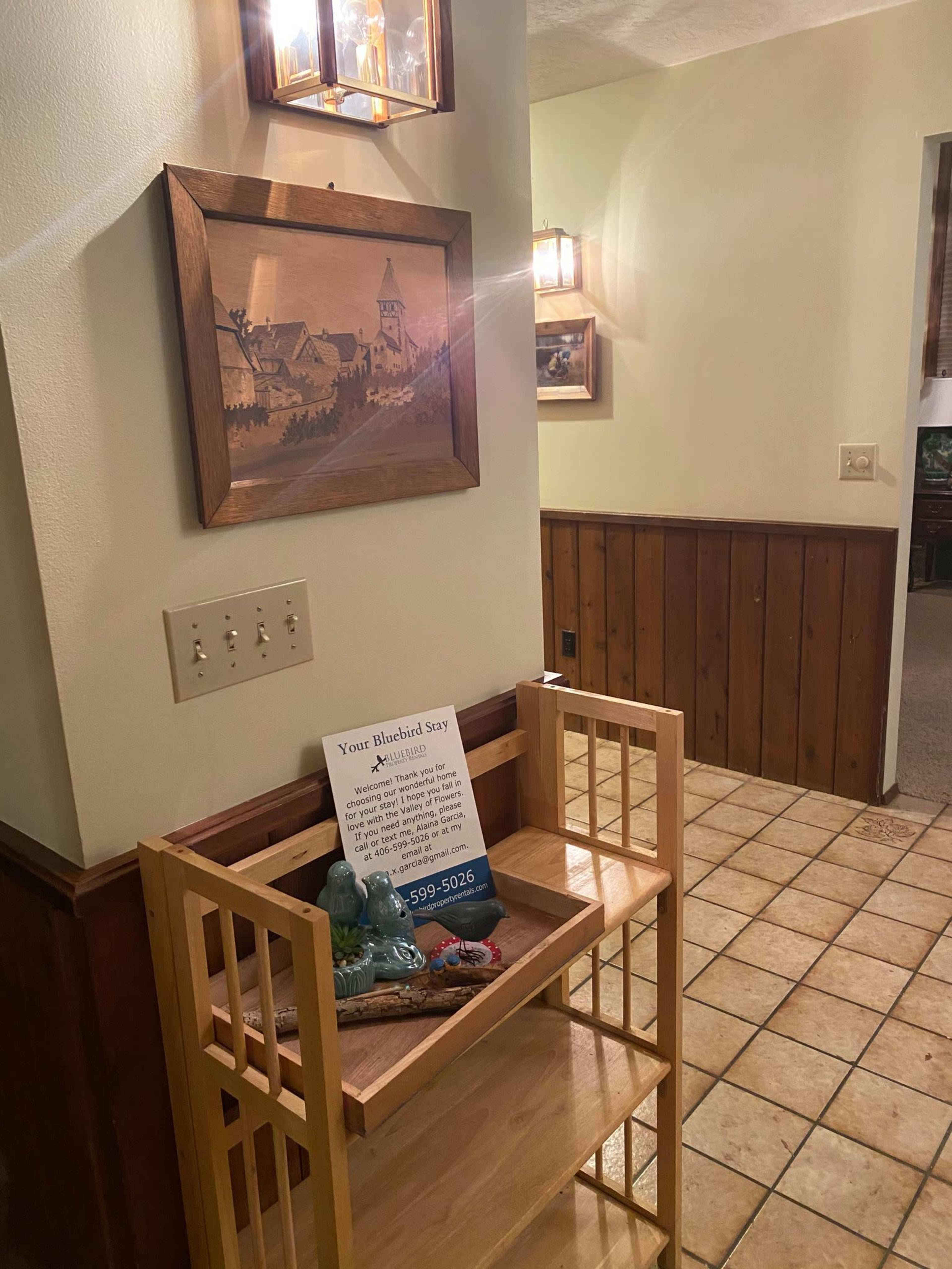 A wooden side table with a sign and decorative items is positioned near a wall with a picture and lights, leading to a hallway with wood paneling.