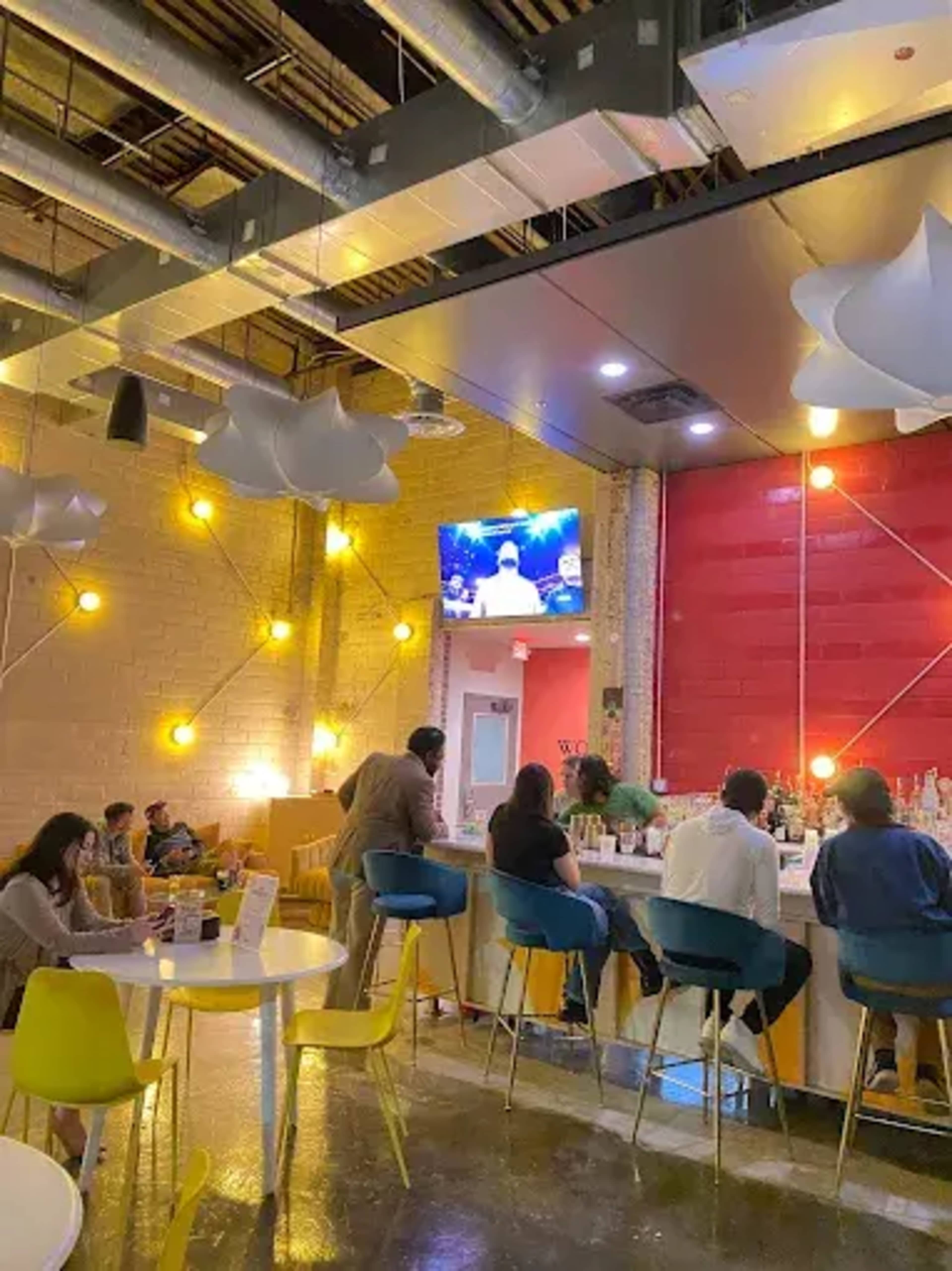 A modern bar area with patrons seated at high-top tables and a large screen displaying a show in the background.