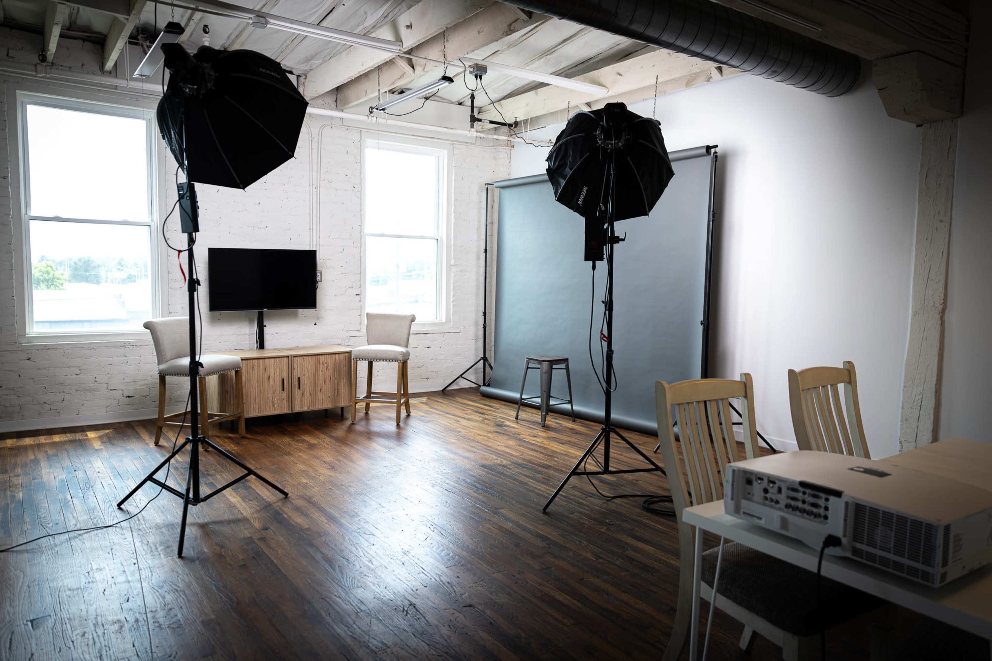 The image shows a bright studio space equipped with three light stands, a gray backdrop, a television on a wooden console, and a table with a projector.