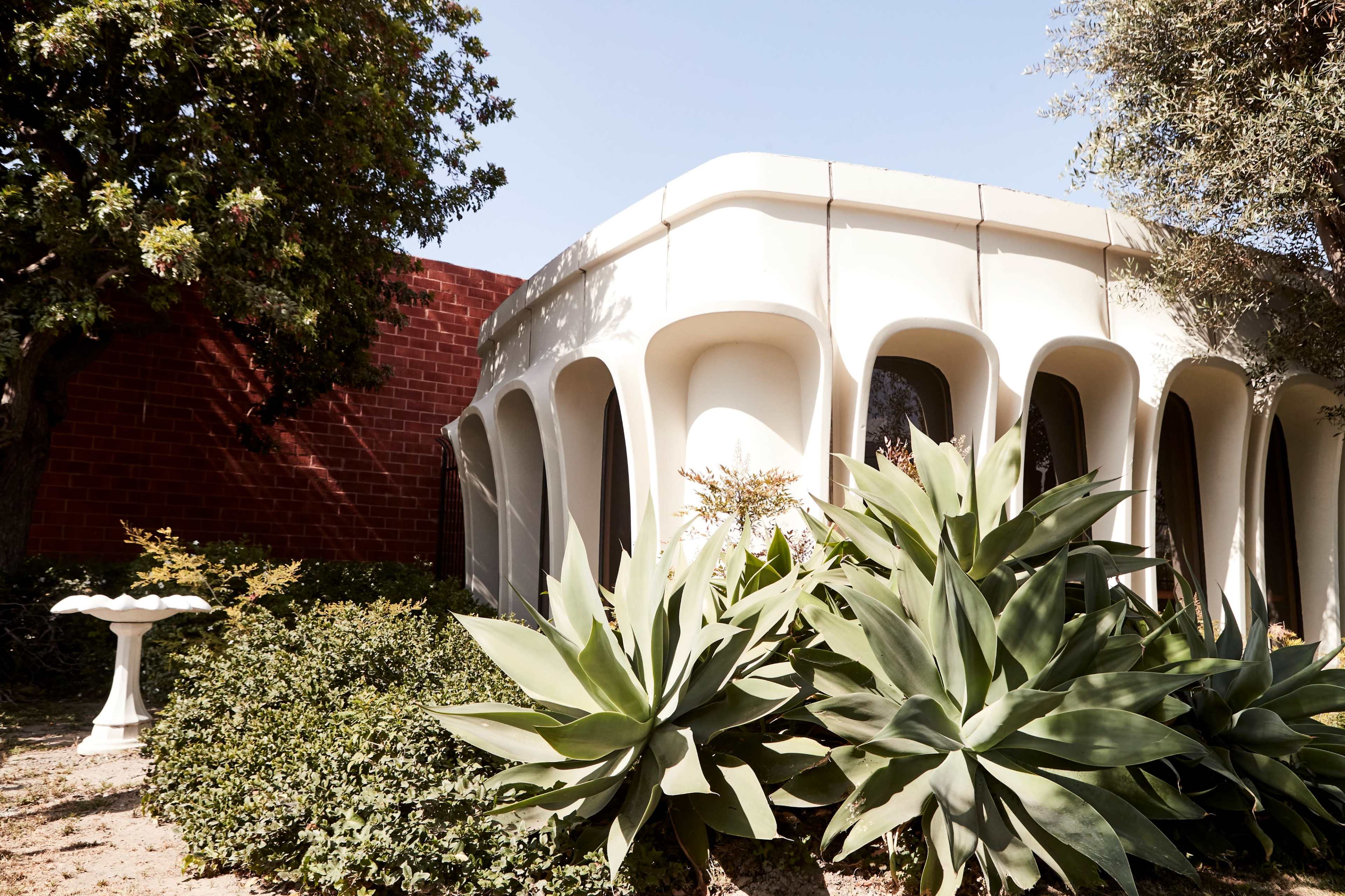 A modern white building with unique architectural features surrounded by lush greenery and a birdbath in the foreground.