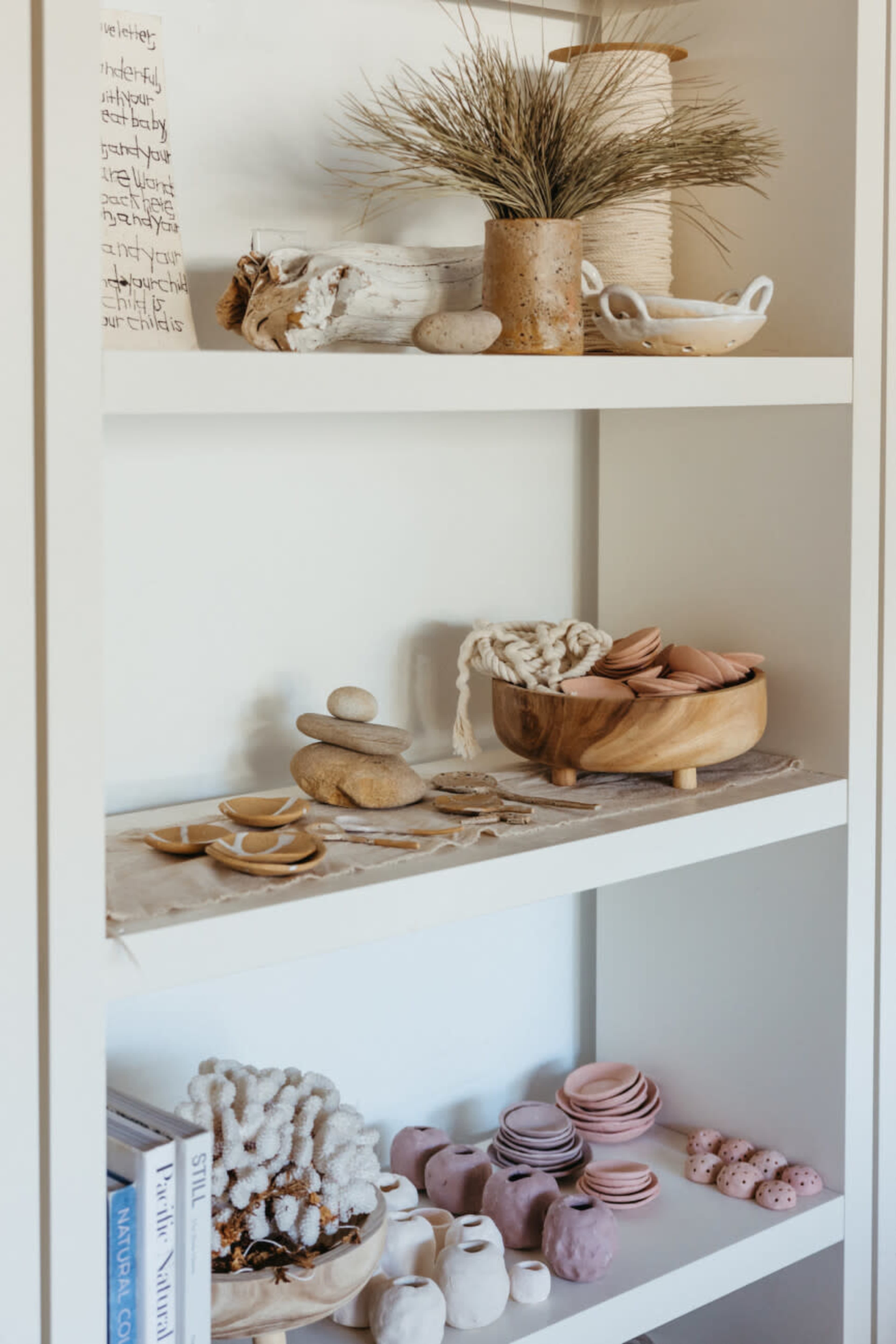 The image shows a curated shelf displaying various decorative items, including ceramics, natural elements, and handmade crafts arranged in a minimalist style.