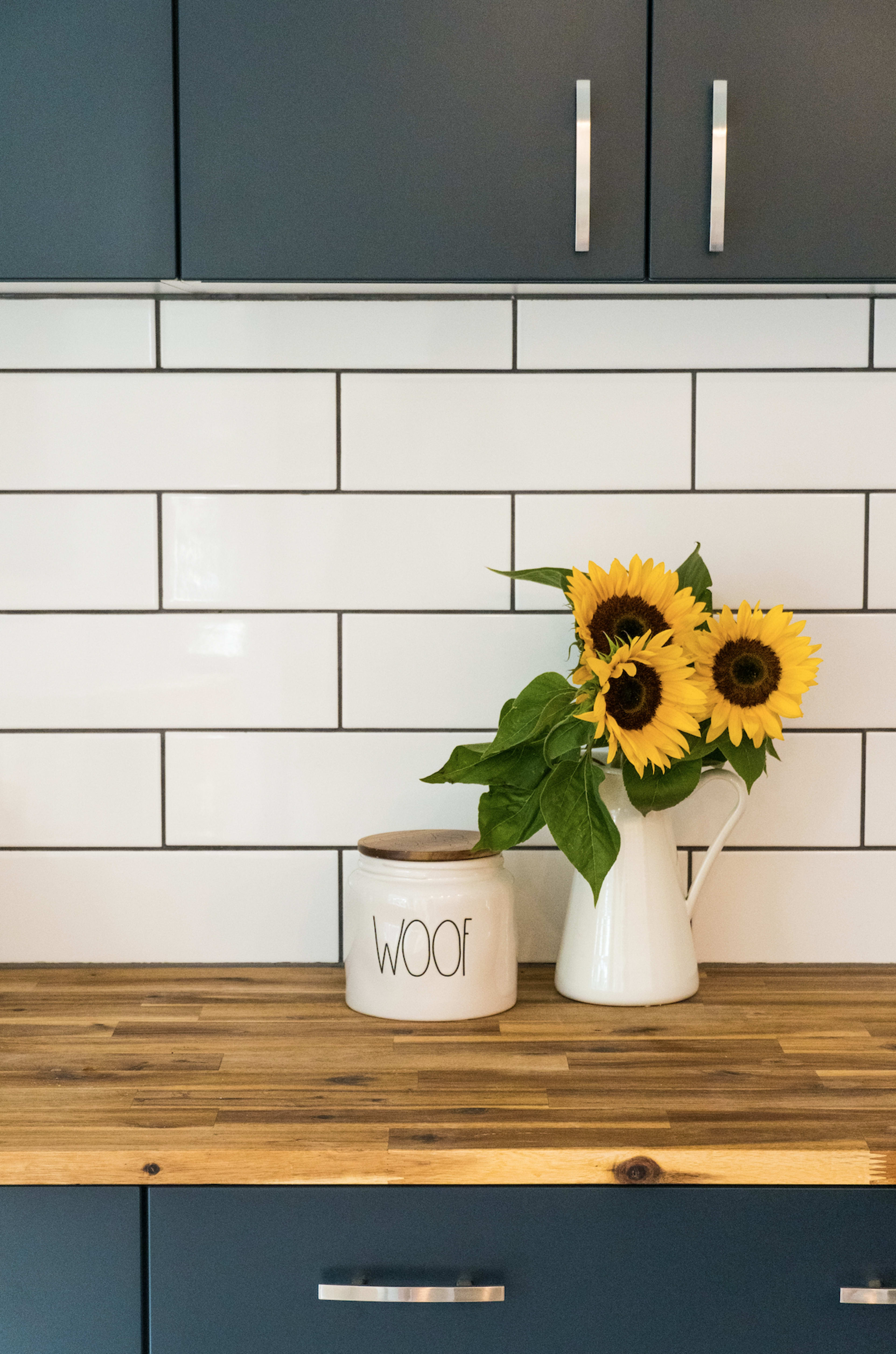 A kitchen counter with a wooden surface featuring a white canister labeled "WOOF" and a white pitcher holding sunflowers, against a backdrop of navy cabinets and white tiled backsplash.