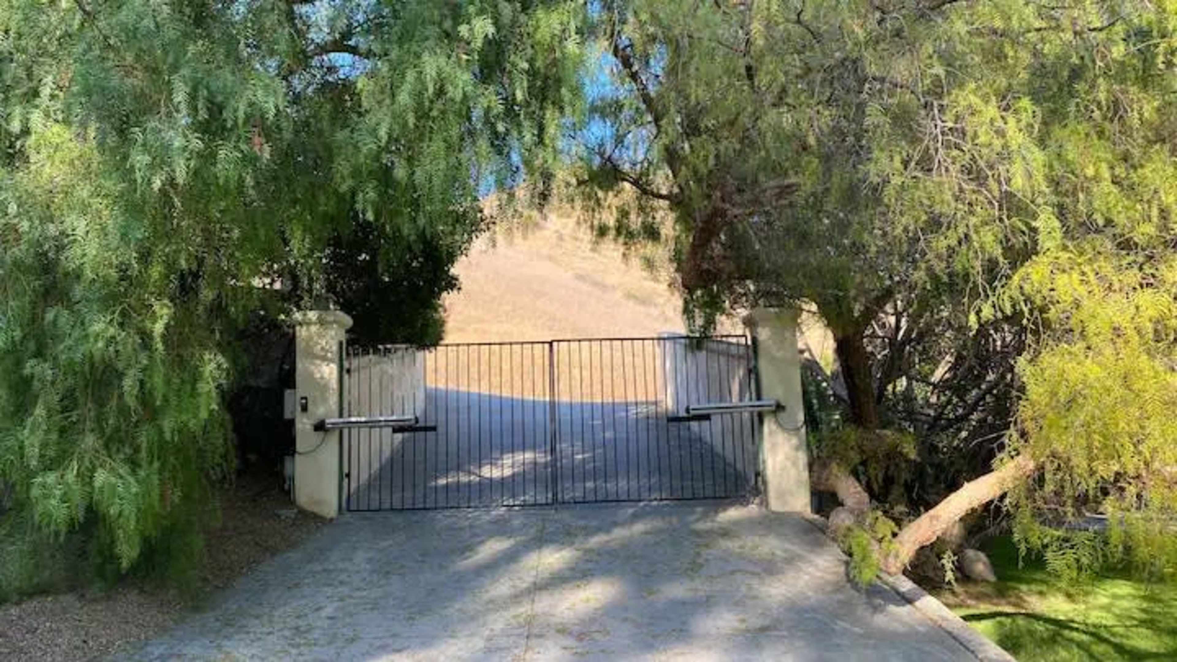 A gated entrance is flanked by trees leading to a sloped driveway.