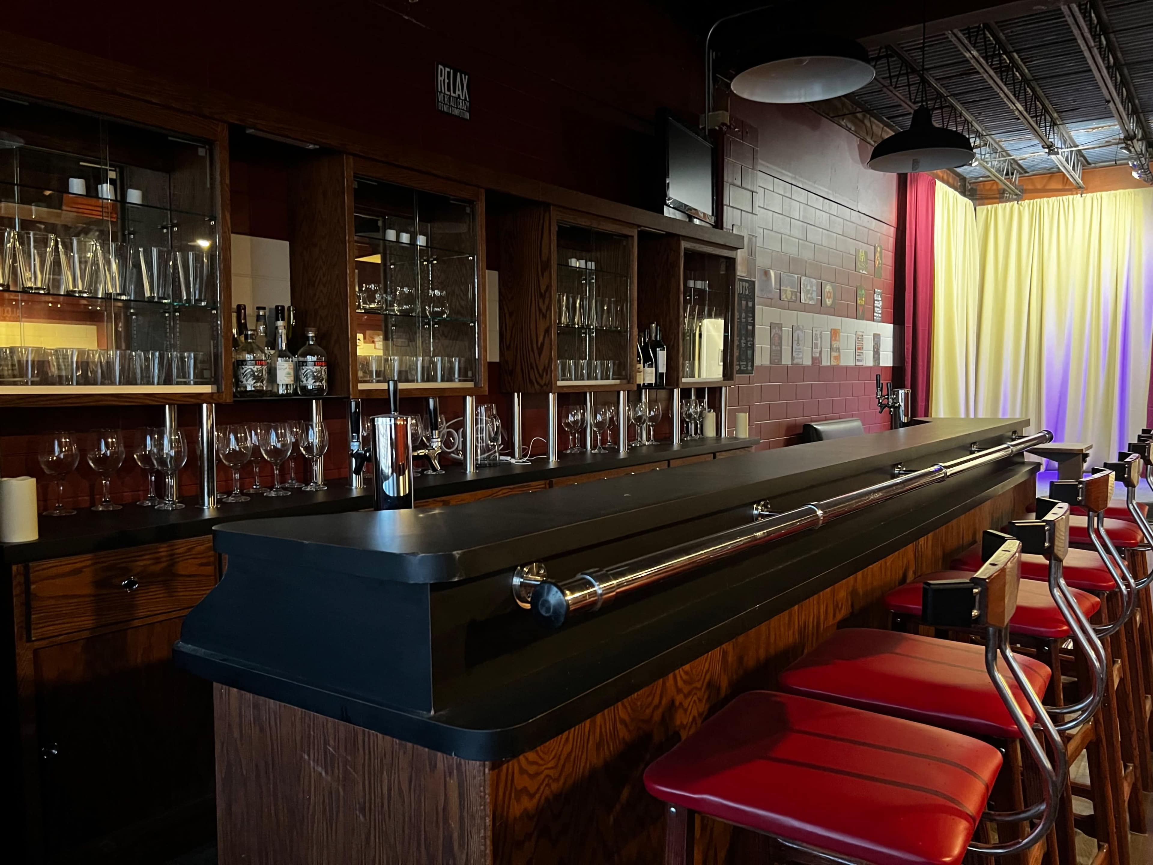 A wooden bar with a long countertop, adorned with glasses and bottles, accompanied by red barstools against a backdrop of relaxed lighting and draped curtains.