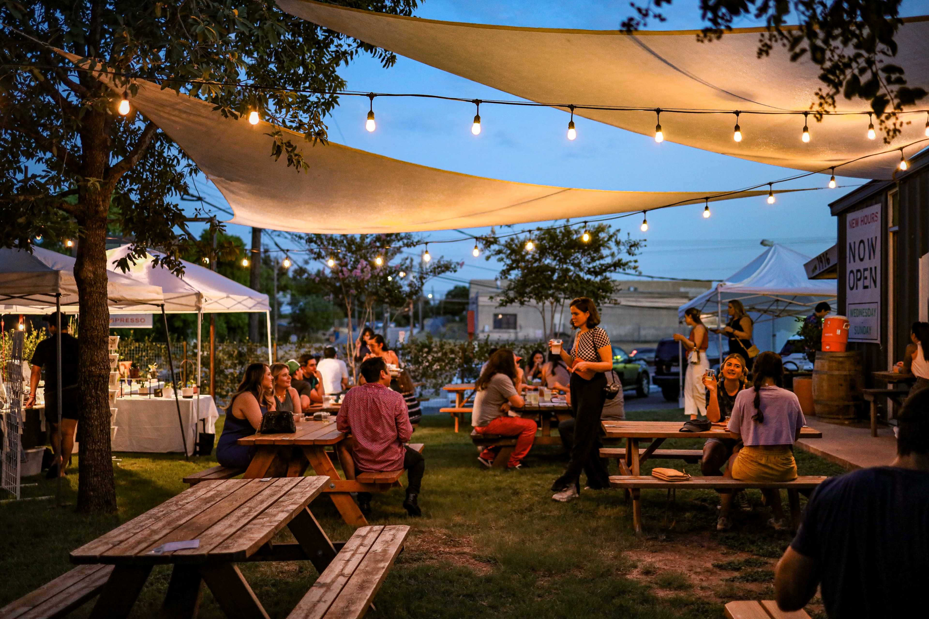 A lively outdoor market features people socializing at wooden picnic tables under string lights and canopies at dusk.