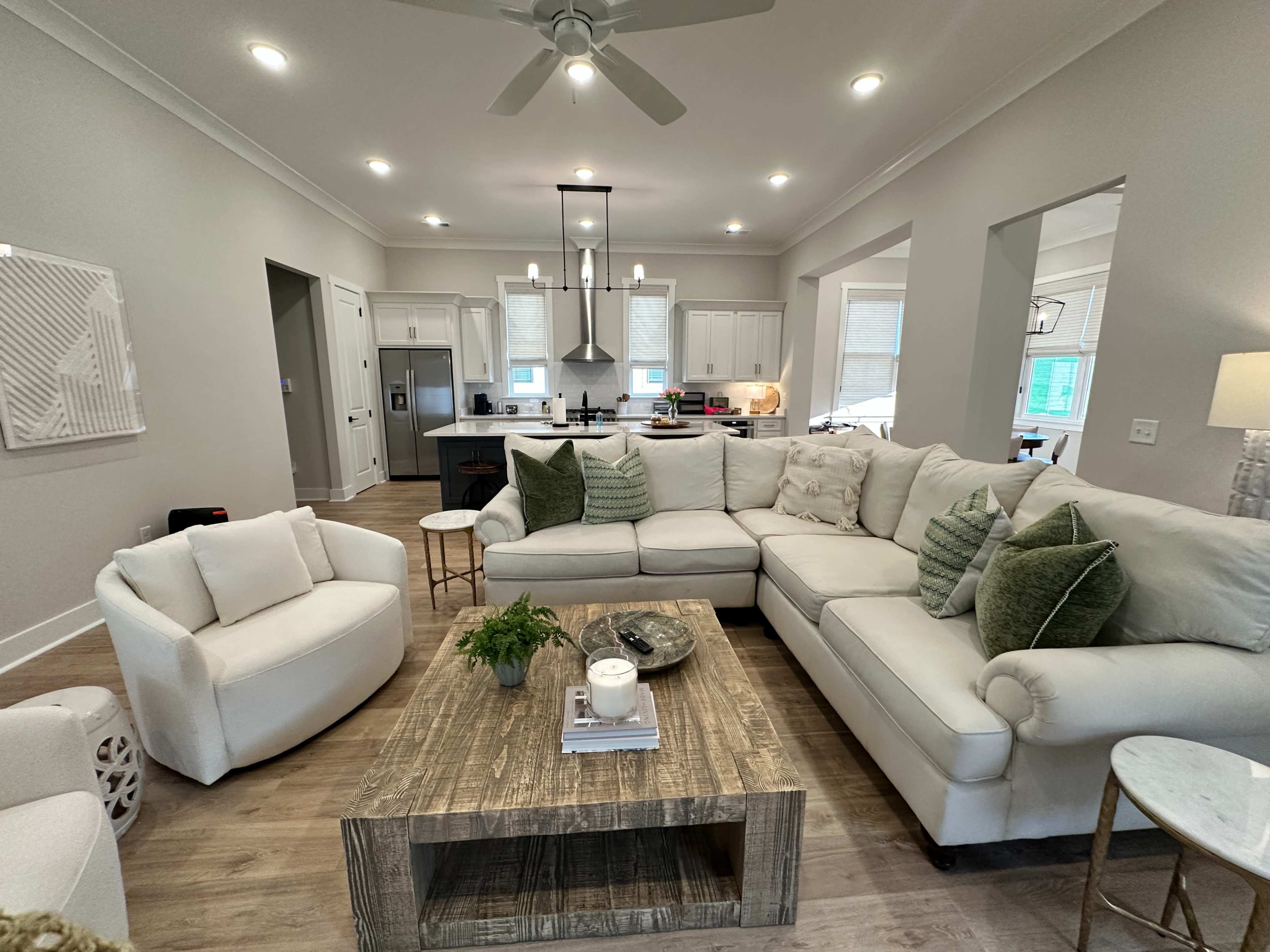 A spacious living room features a large white sectional sofa arranged around a wooden coffee table, with a view of a modern kitchen in the background.