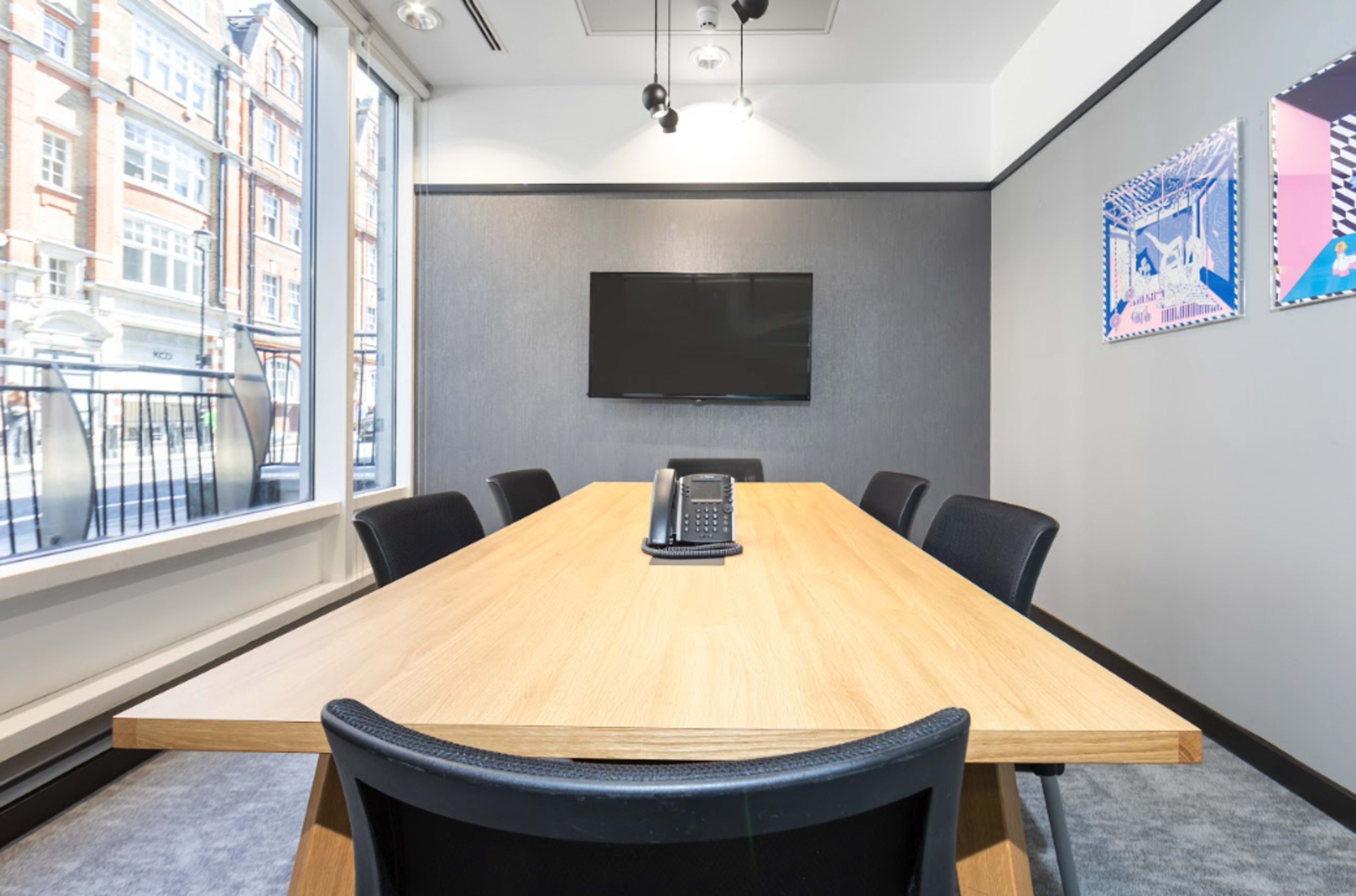 The image shows a modern conference room with a long wooden table, black chairs, a wall-mounted TV, and large windows letting in natural light.