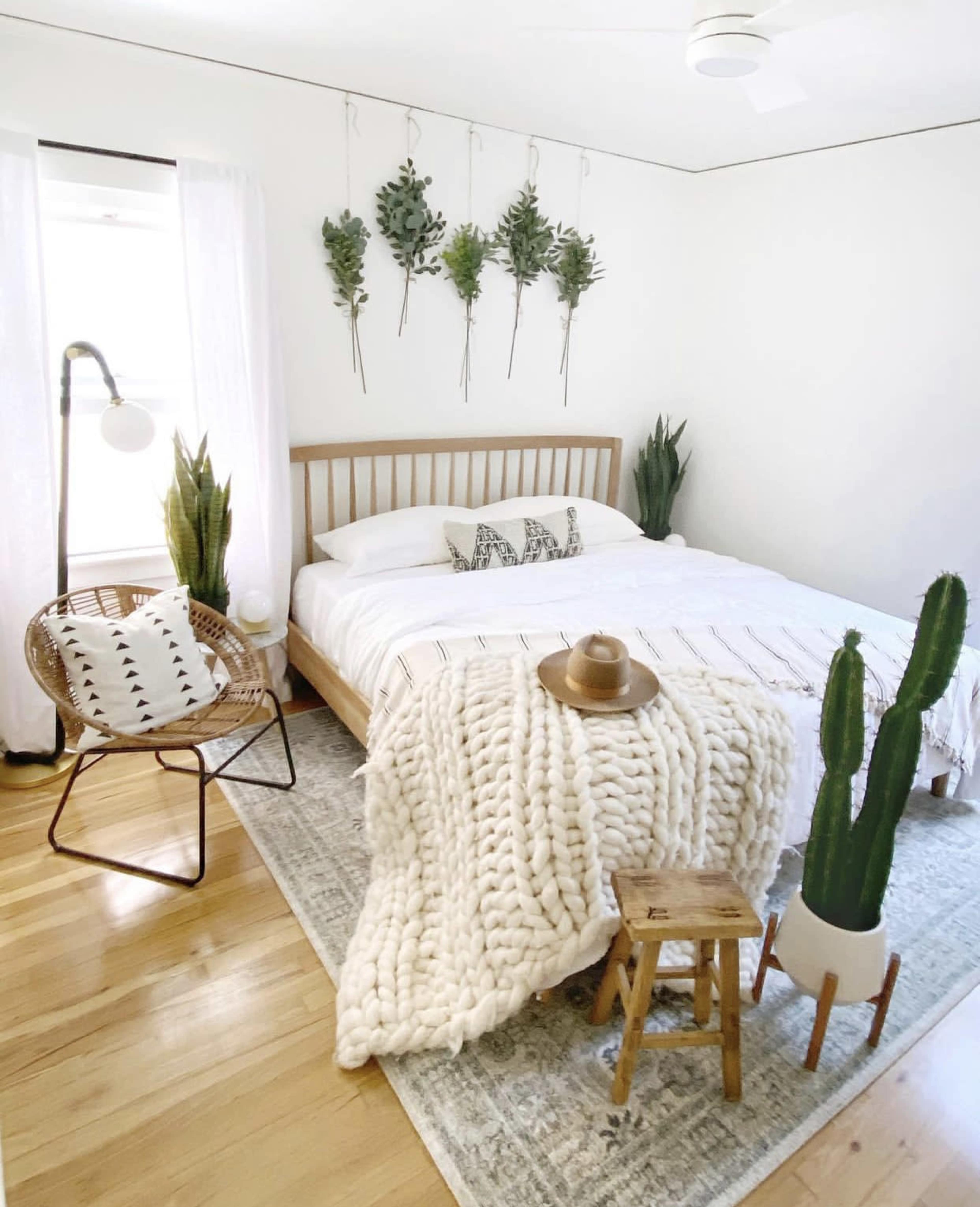 A minimalist bedroom with a bed, a woven chair, and potted plants, featuring a cozy blanket and decorative accents on the walls.