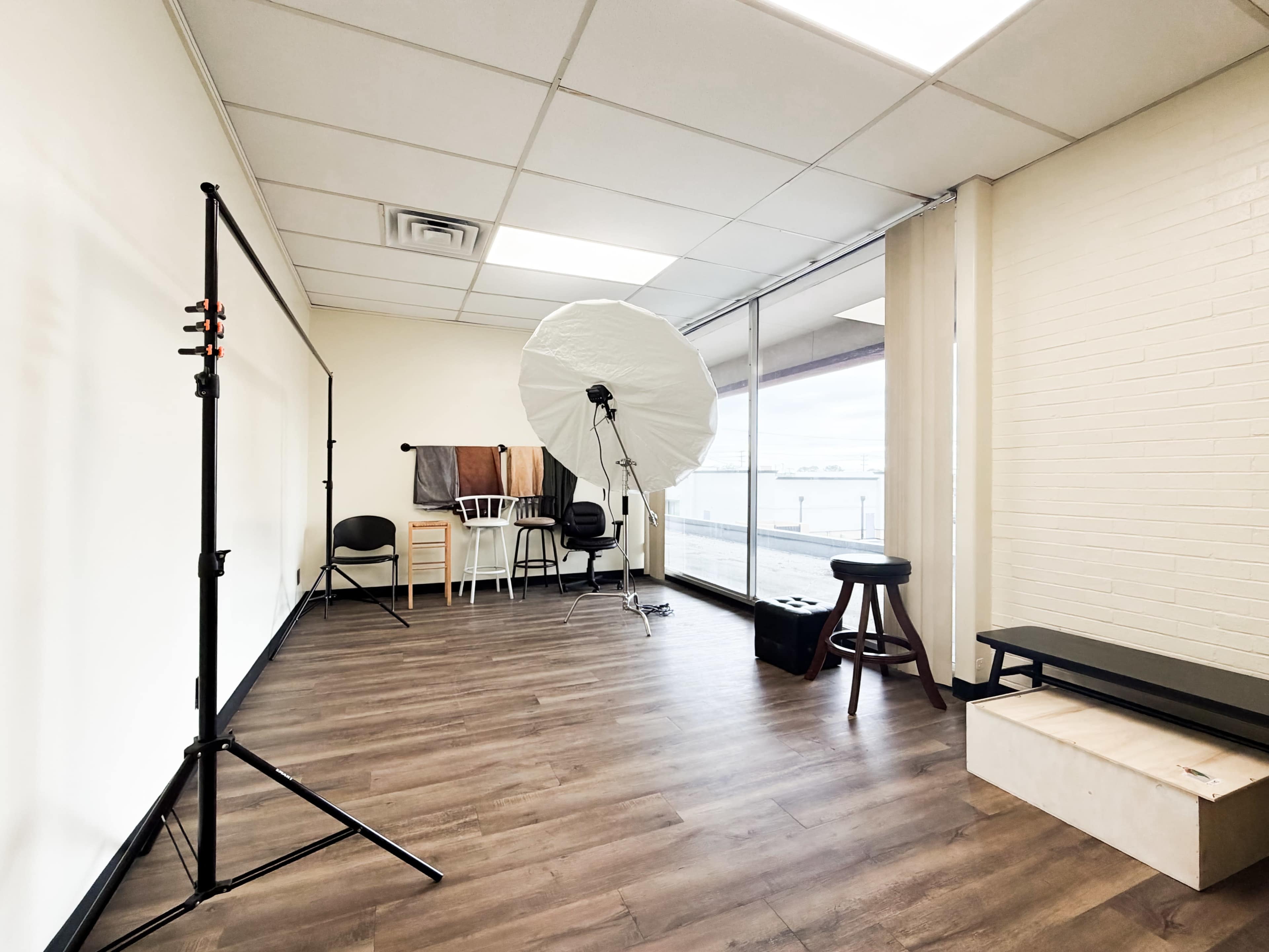 The image shows a photography studio with a large softbox light, a stool, a black bench, a few chairs, and a window letting in natural light.
