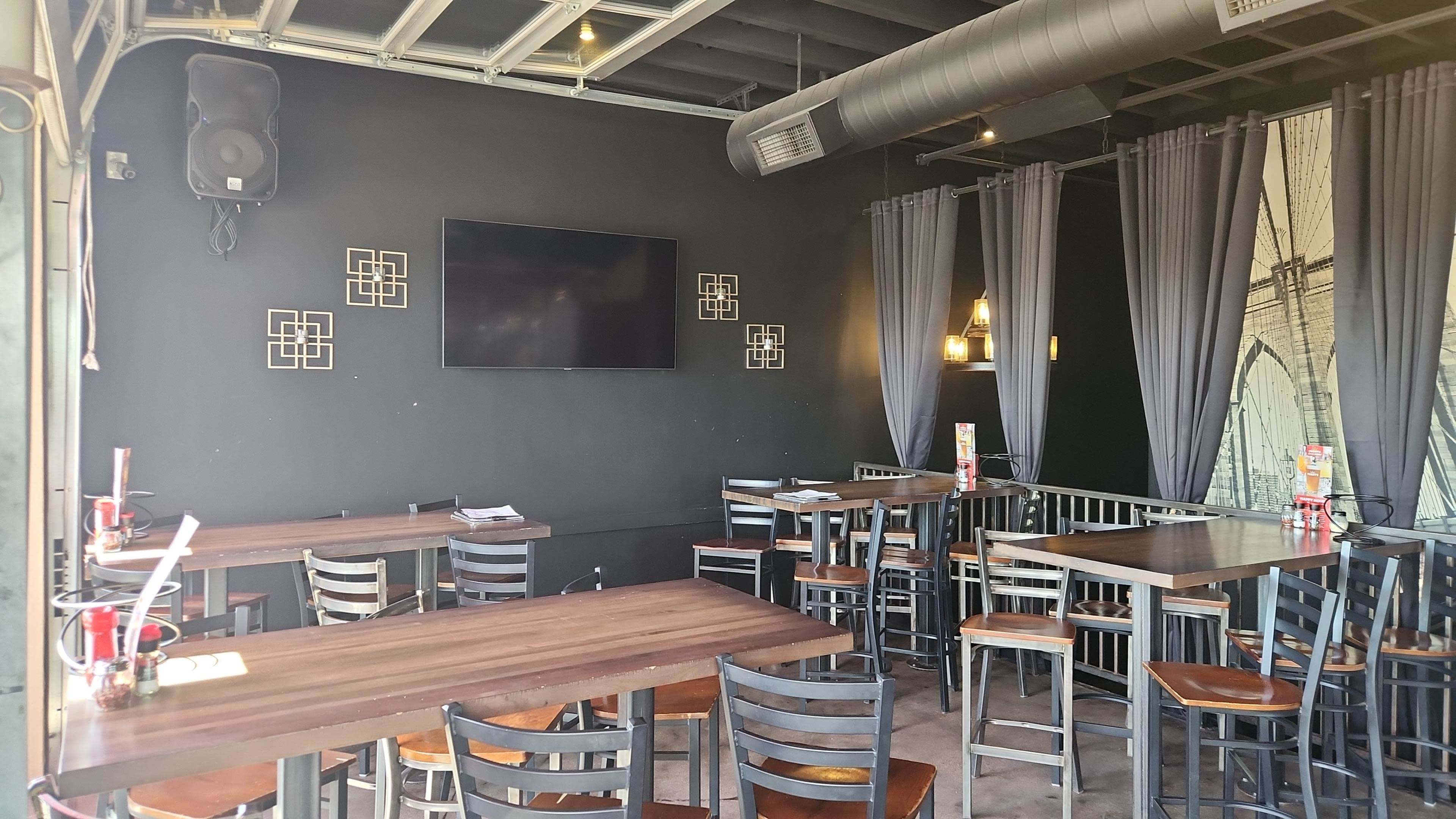 The image shows an indoor dining area of a restaurant with wooden tables and black chairs, featuring a large wall-mounted TV and gray curtains.
