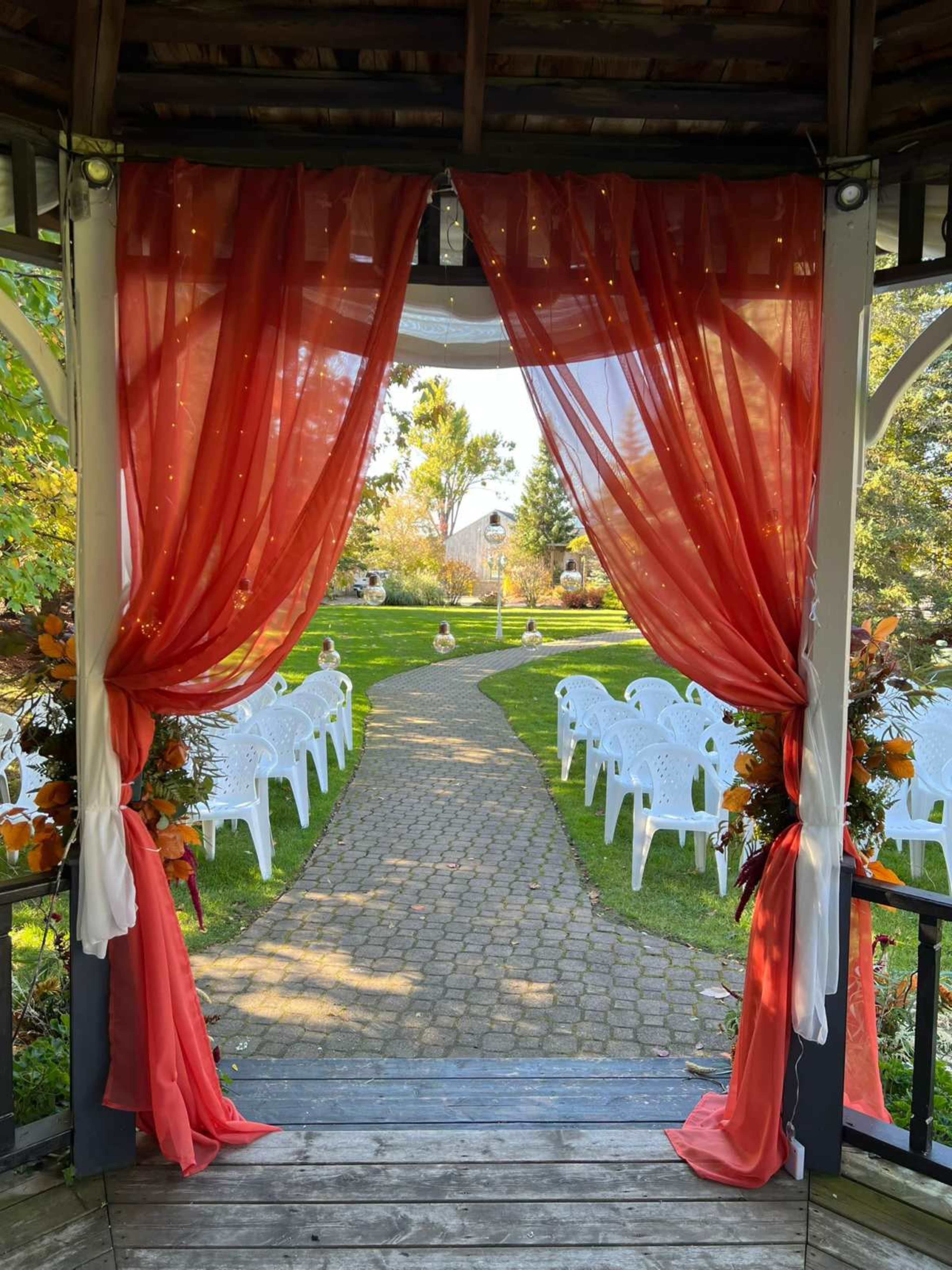 The image shows a gazebo with flowing red curtains framing a pathway leading to a landscaped garden with white chairs arranged for an outdoor event.