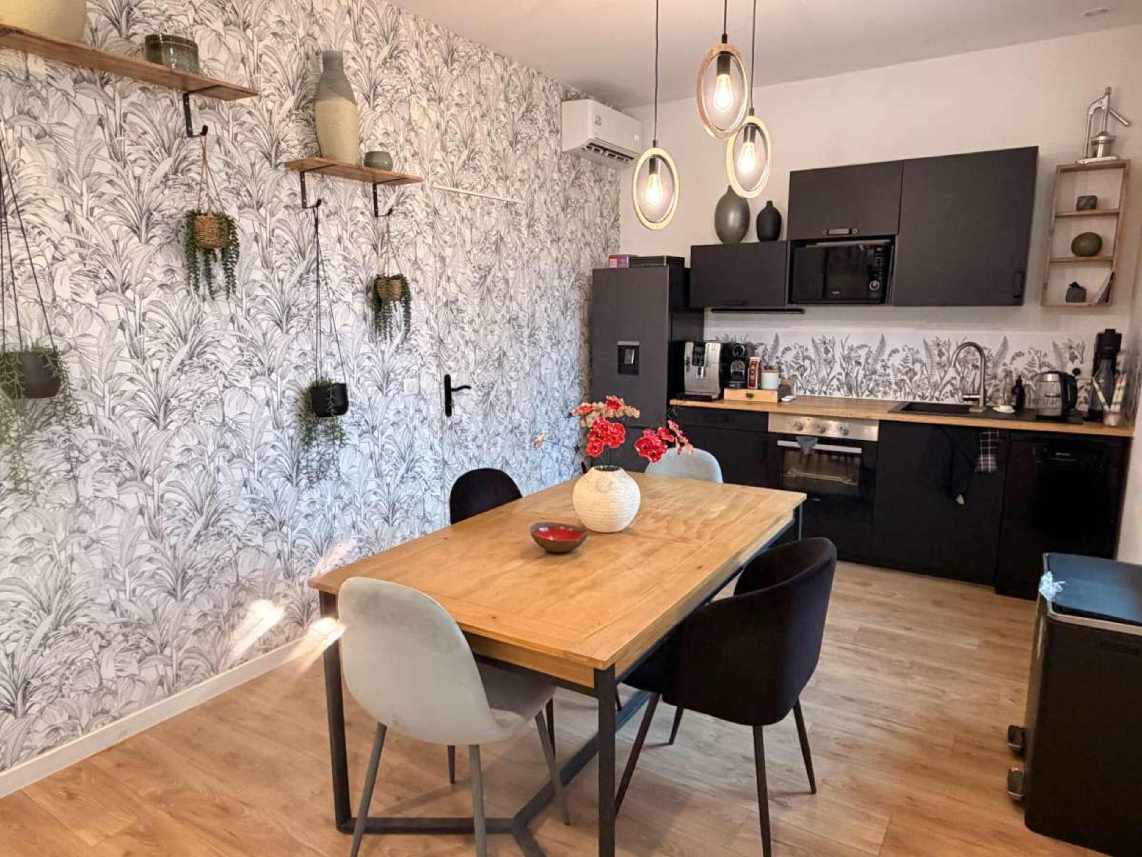 The image shows a dining area with a wooden table surrounded by four chairs, set against a patterned wall and a kitchen featuring dark cabinetry and various kitchen appliances.