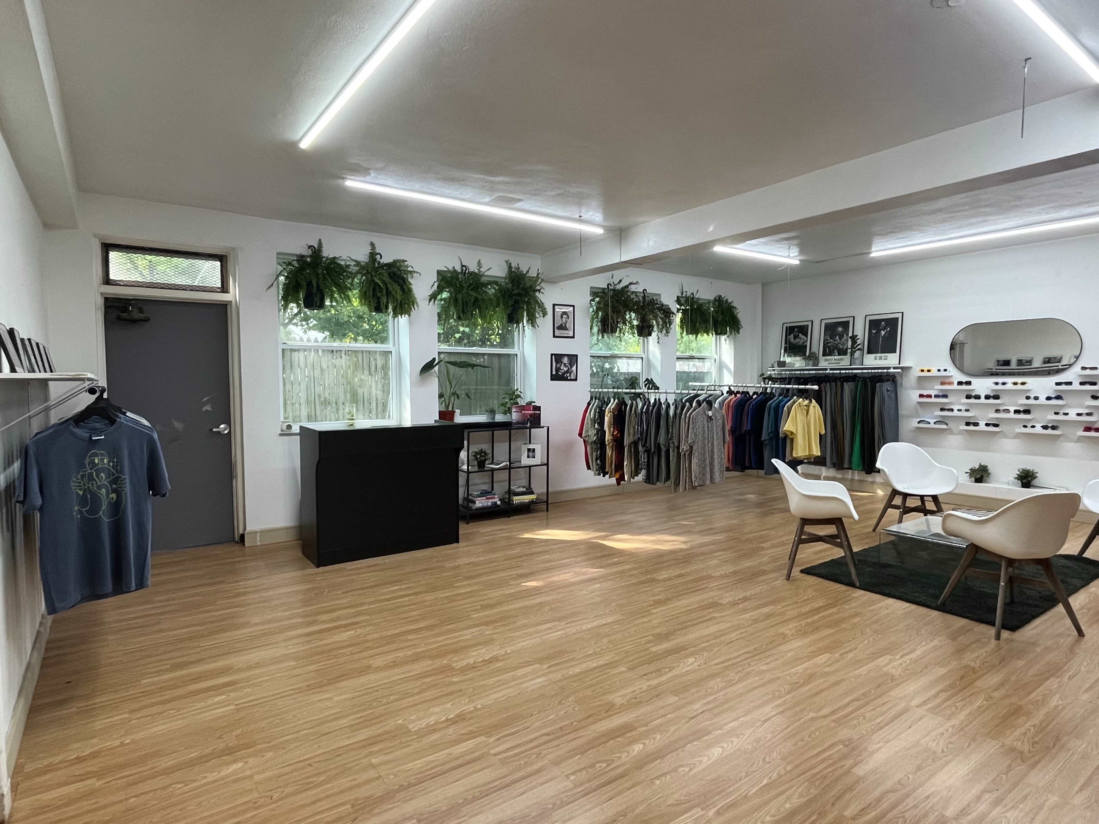 A clothing boutique with a minimalist interior, featuring racks of apparel, a reception counter, and seating arranged on wooden floors.