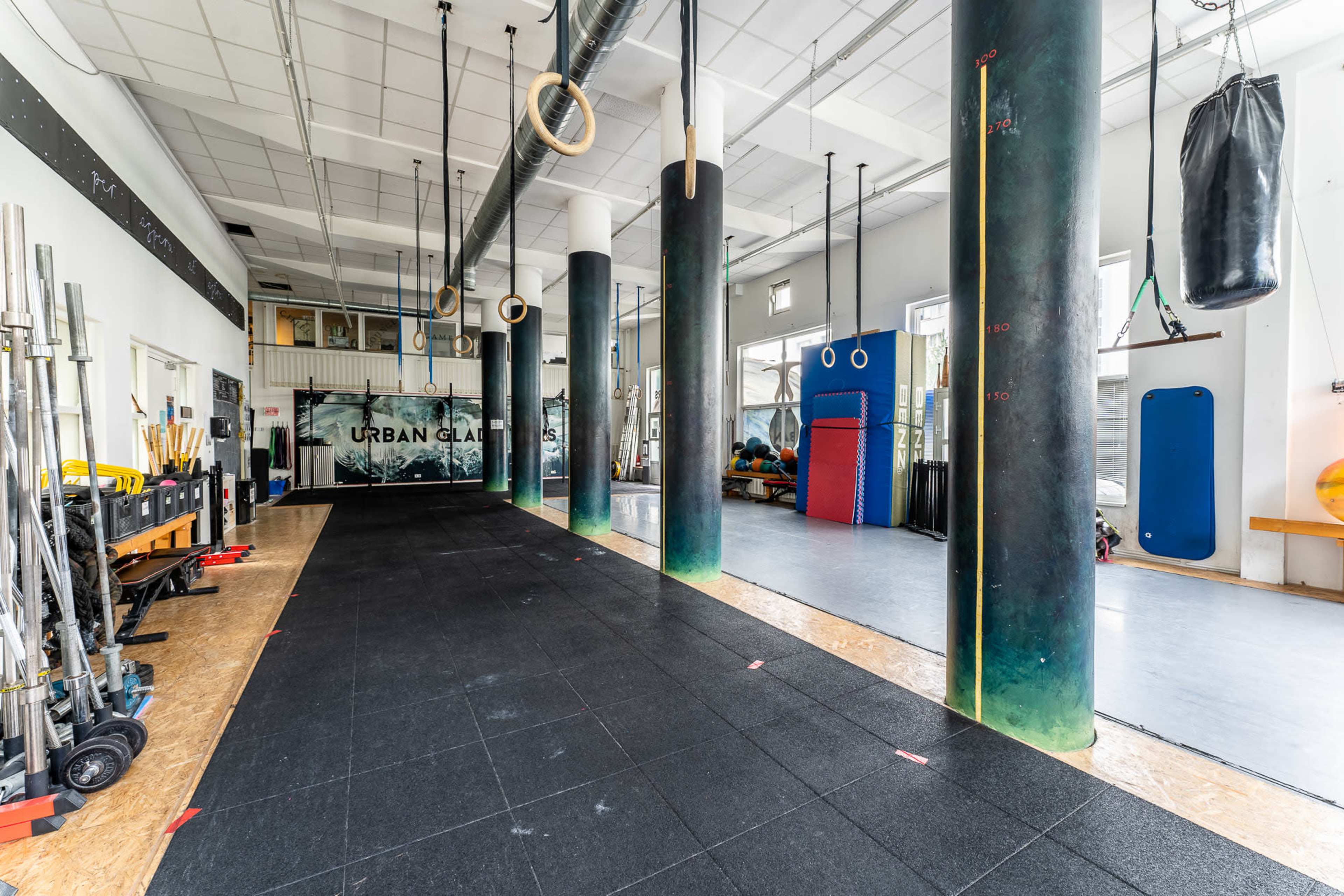 A spacious gym with rubber flooring, large columns, fitness equipment along one side, and various training apparatuses in the background.