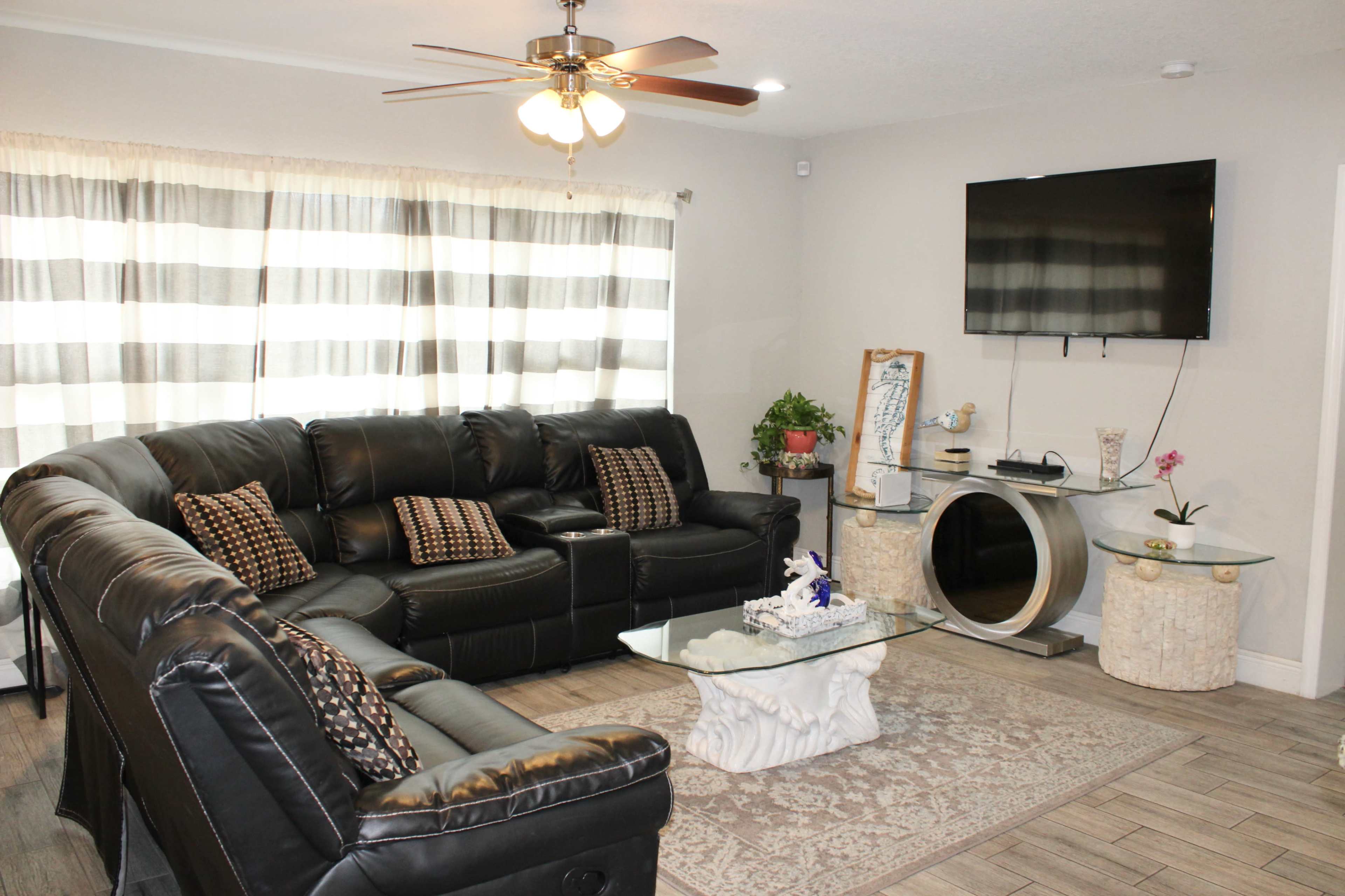 A living room with a black leather sectional sofa, a glass coffee table on a patterned rug, and a wall-mounted television.