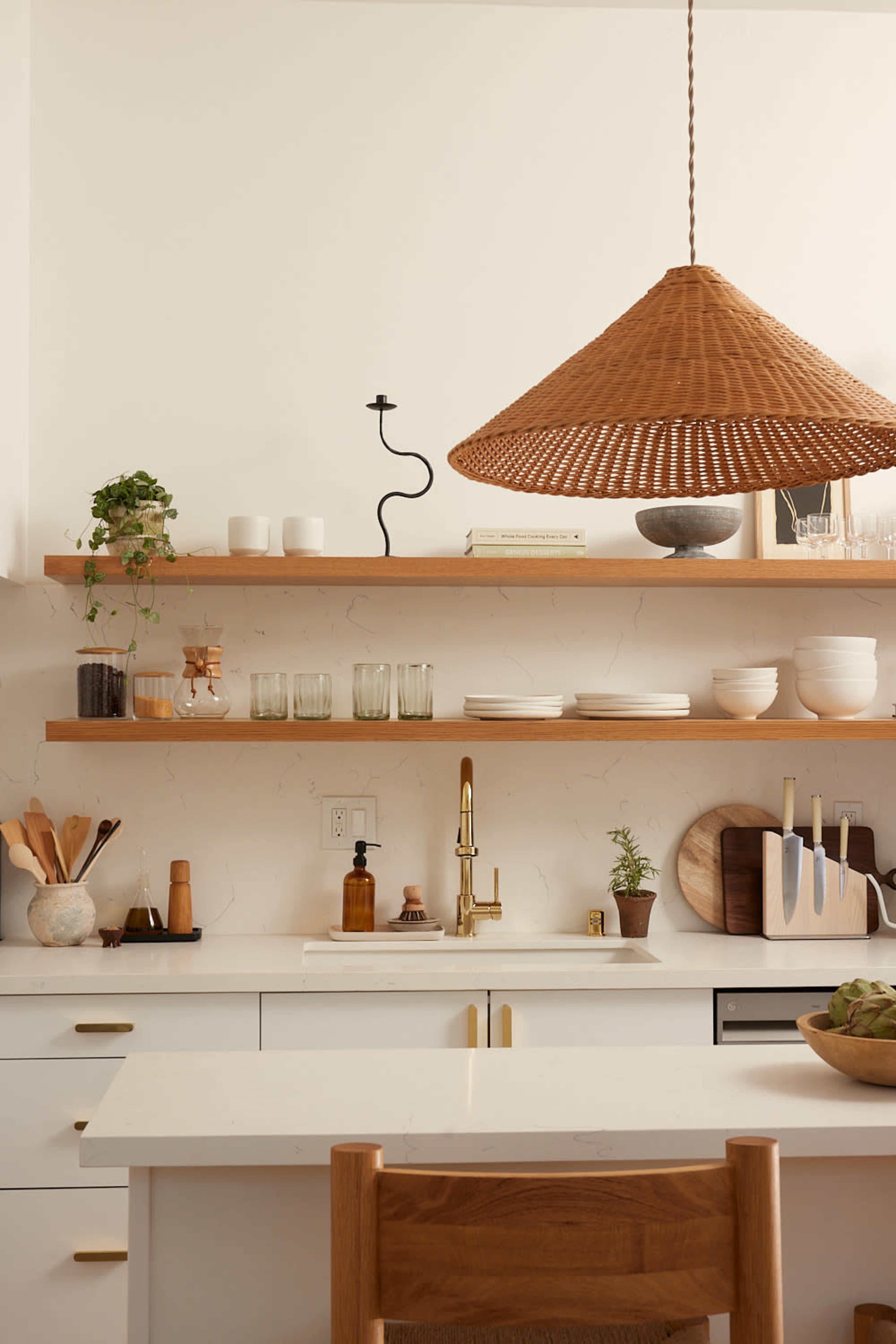 A modern kitchen features open shelving with various dishes, glassware, and plants above a clean countertop with a sink and a brass faucet, illuminated by a woven pendant light.