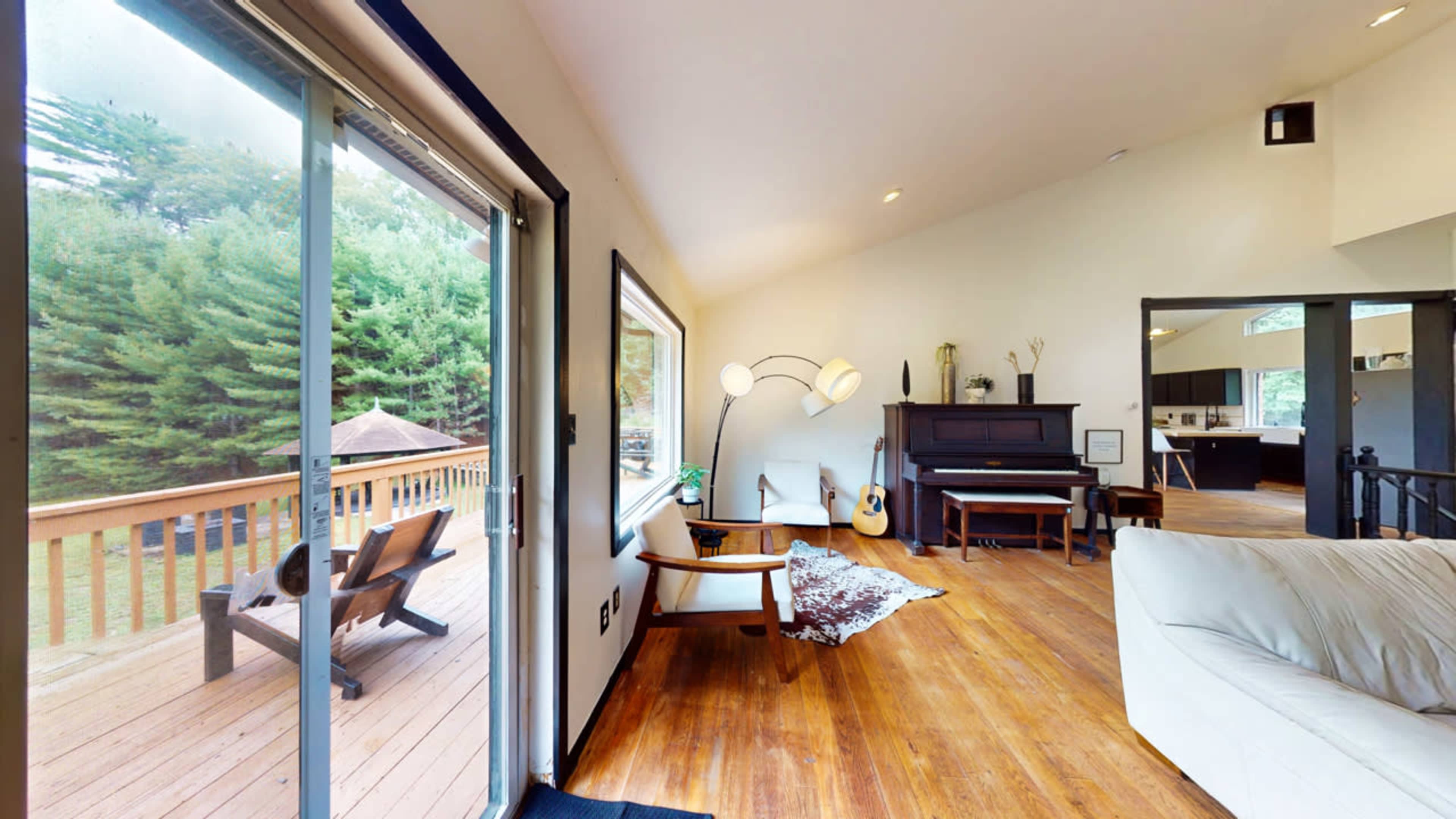 A spacious living room featuring a piano, a guitar, and large windows that overlook a wooden deck with outdoor seating.