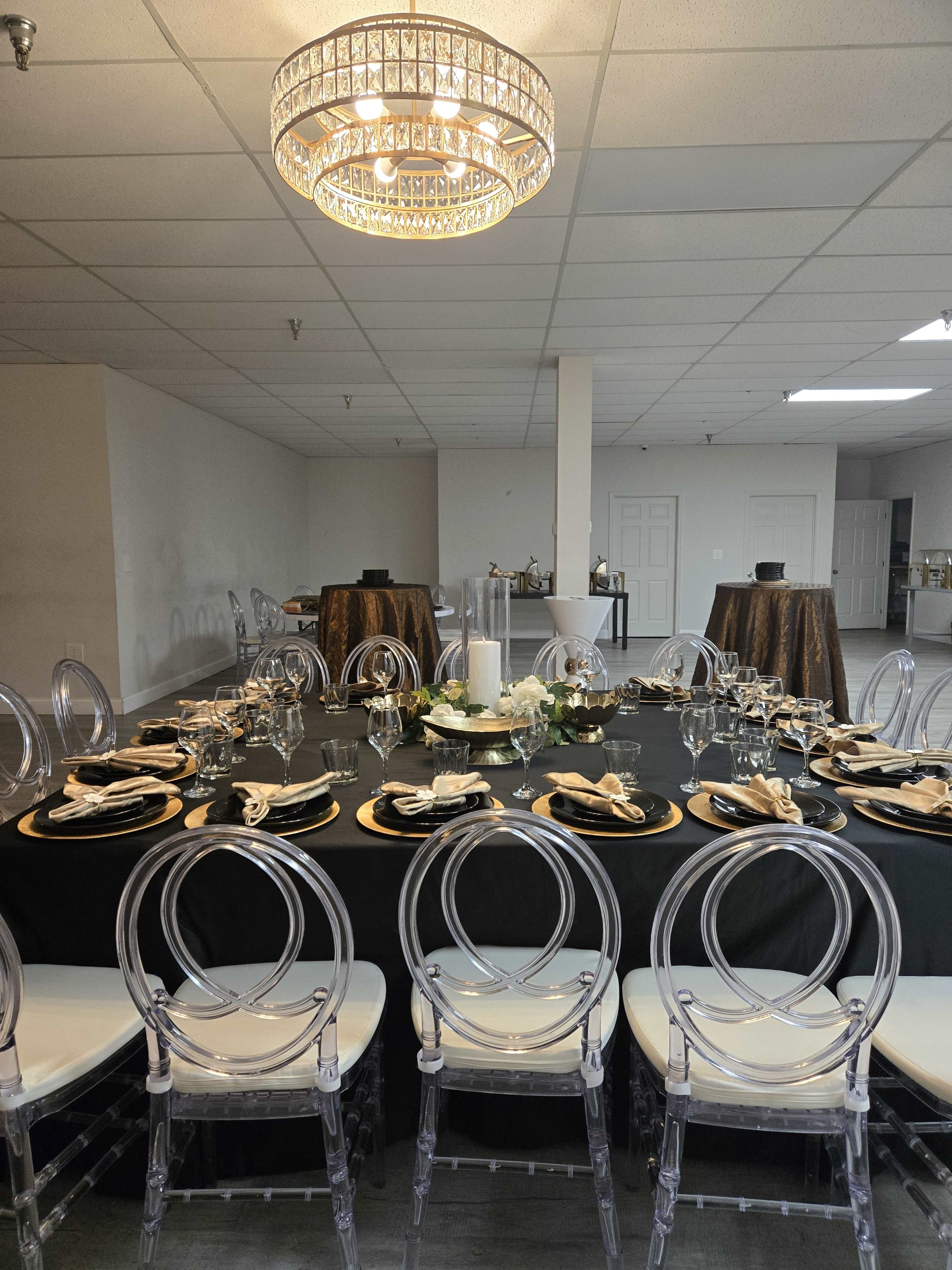 A round dining table is set with elegant place settings and surrounded by clear acrylic chairs, under a sparkling chandelier in a spacious event room.