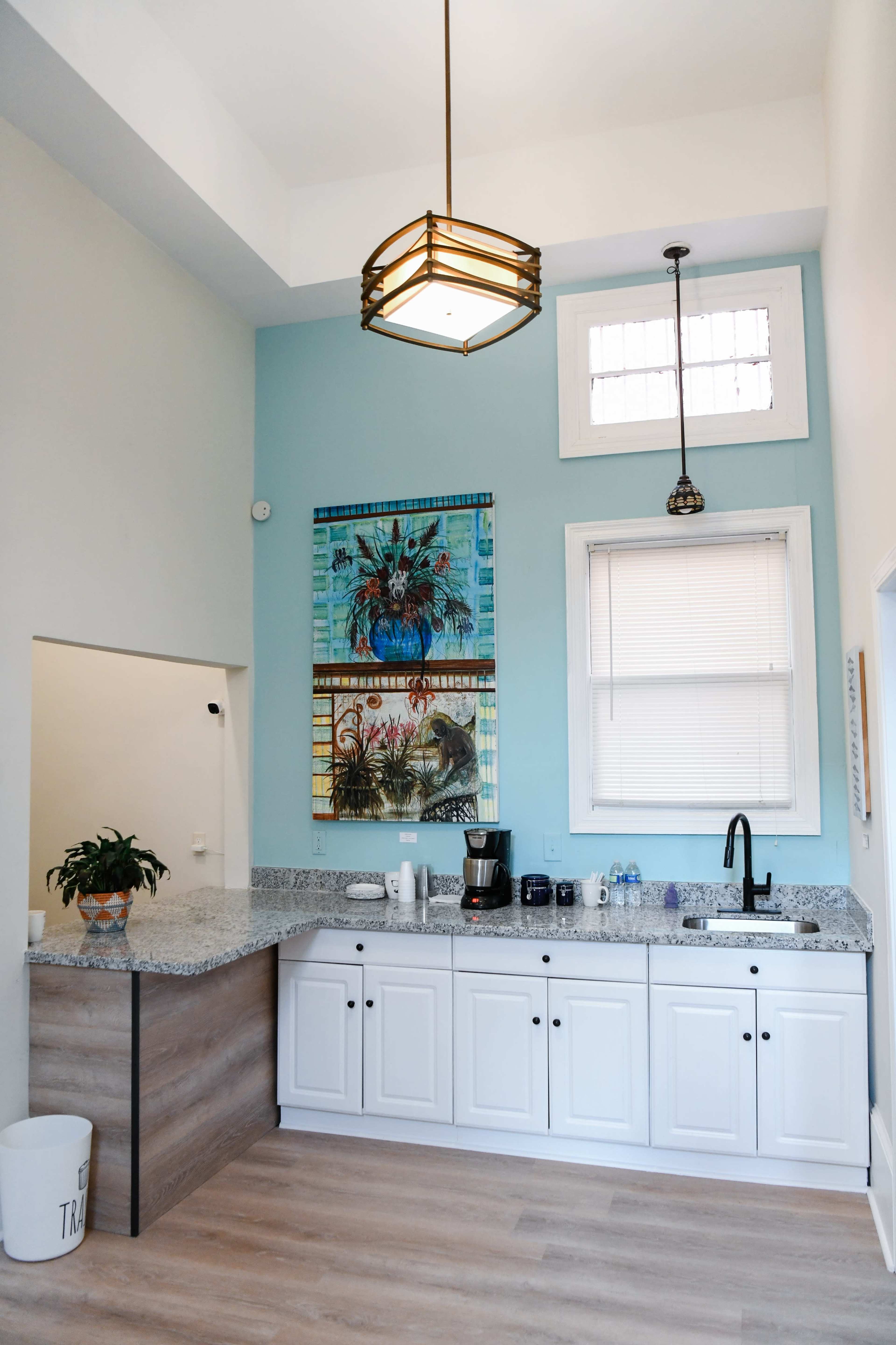 A modern kitchen with a blue wall featuring framed artwork, granite countertops, white cabinets, and a pendant light above the sink.