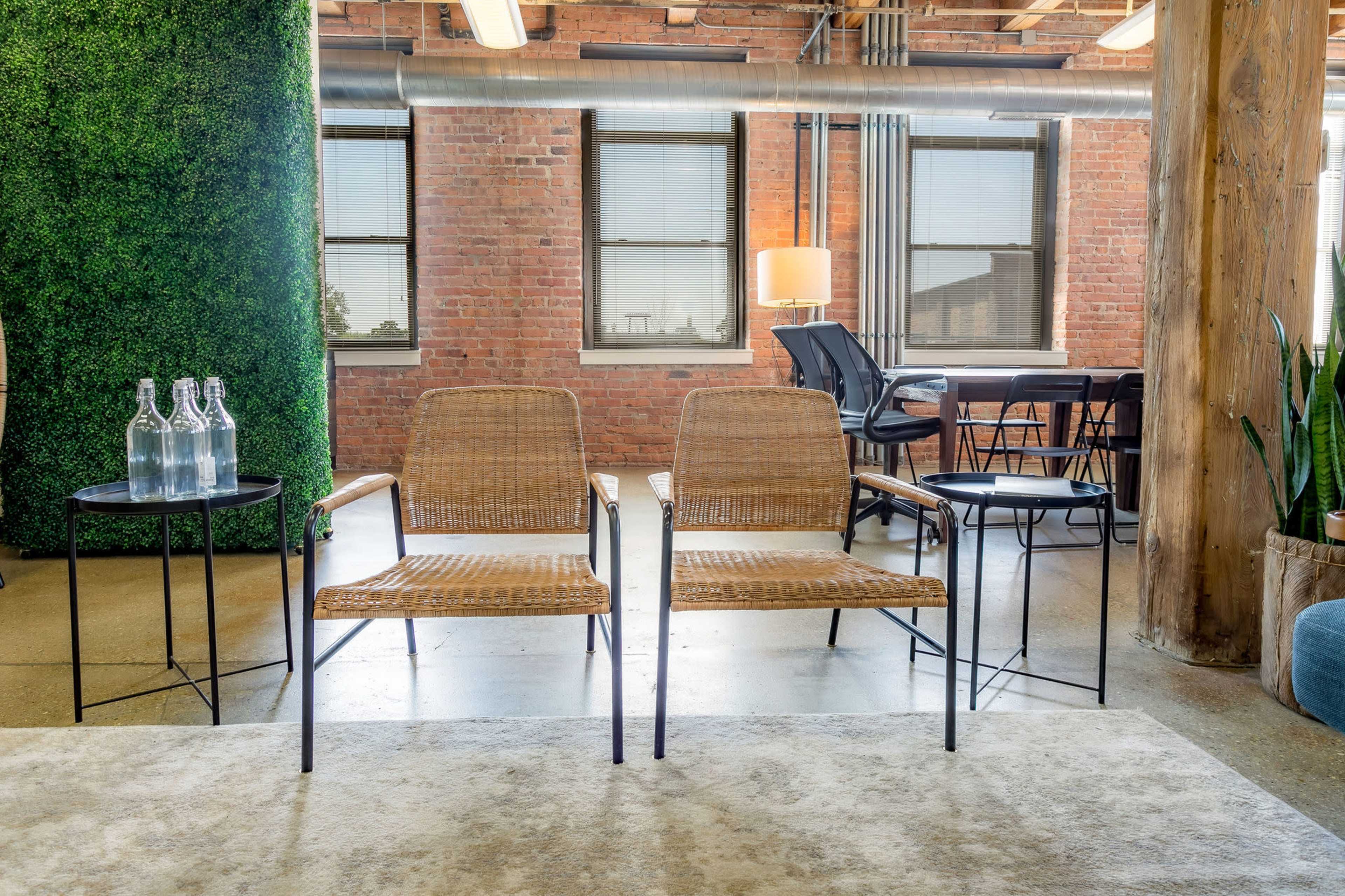 The image features two woven chairs facing each other on a rug, positioned in a modern, open workspace with brick walls and large windows.