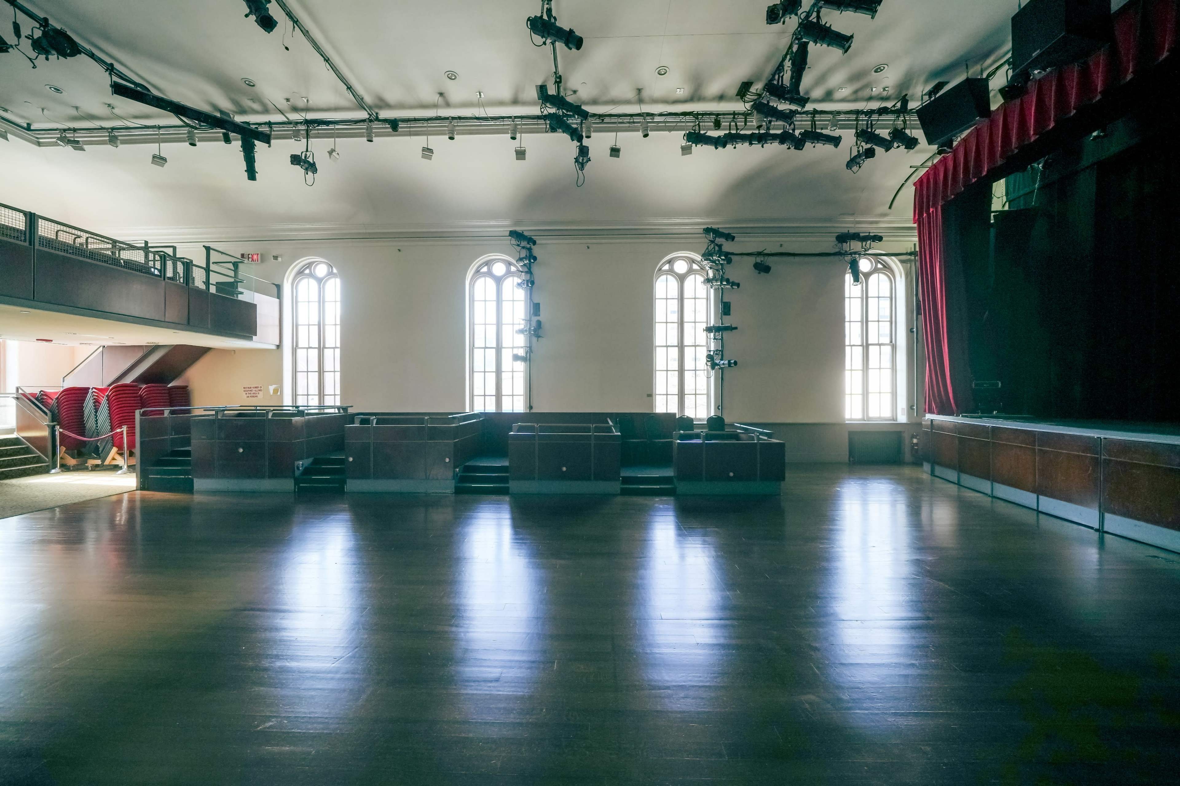 An empty performance space with wooden flooring, large windows, and a stage at the front.