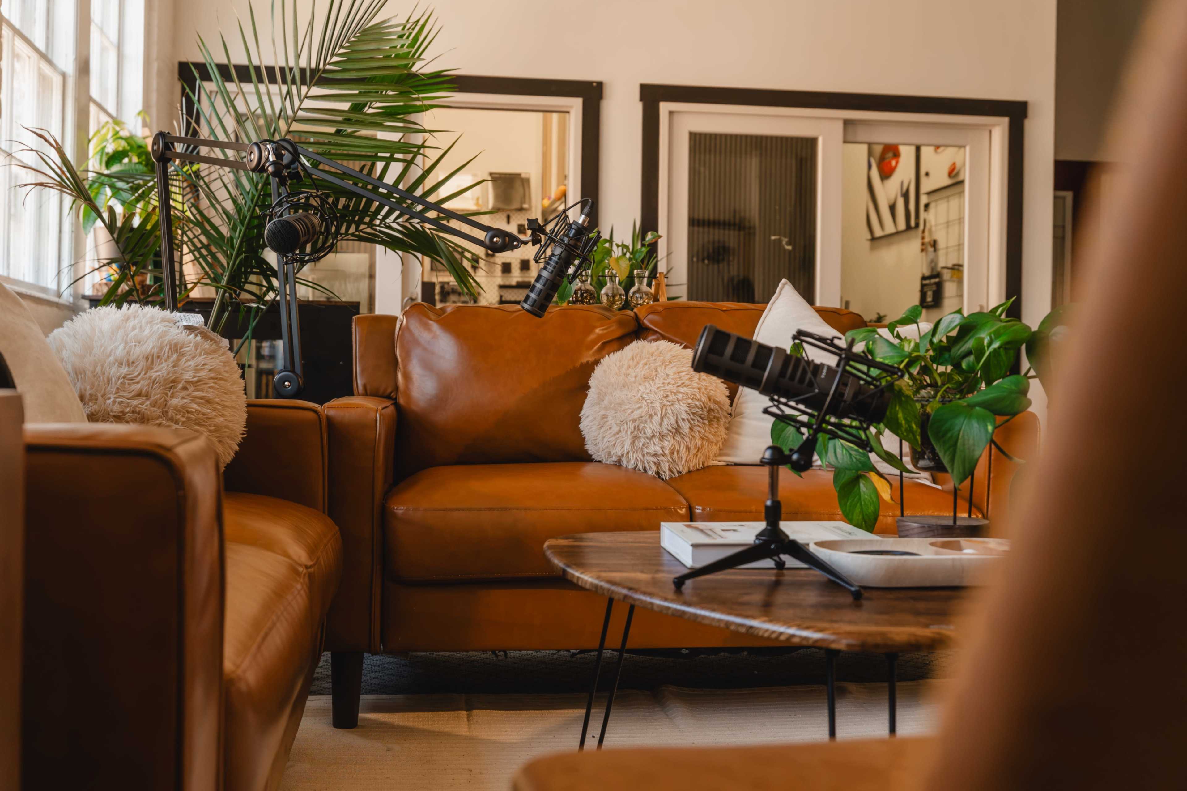 The image shows a cozy seating area with brown leather sofas, a wooden coffee table, and microphones set up for a podcast or recording session, surrounded by houseplants.