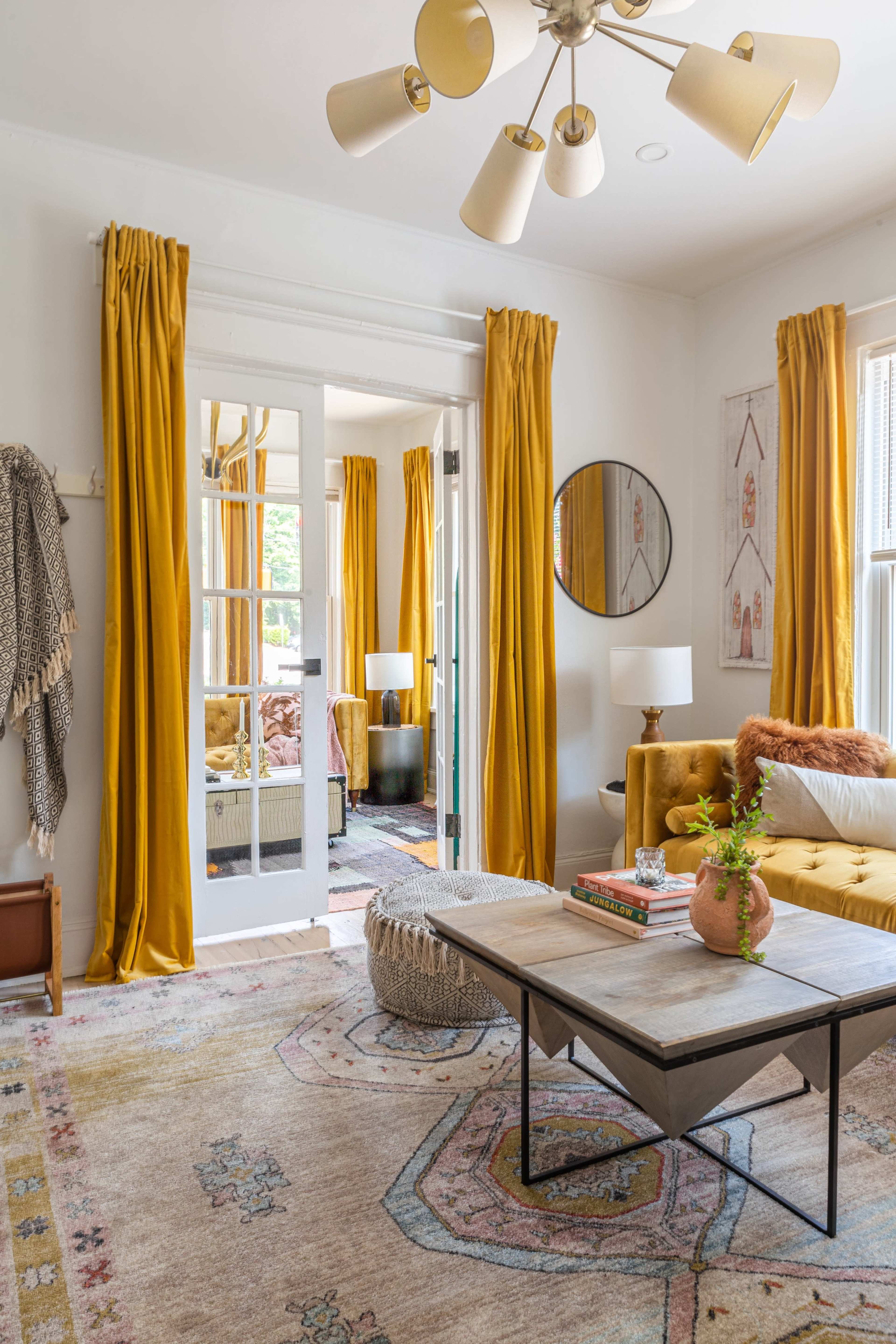 A living room features a coffee table on a patterned rug, surrounded by a mustard yellow sofa, potted plant, and tall curtains.