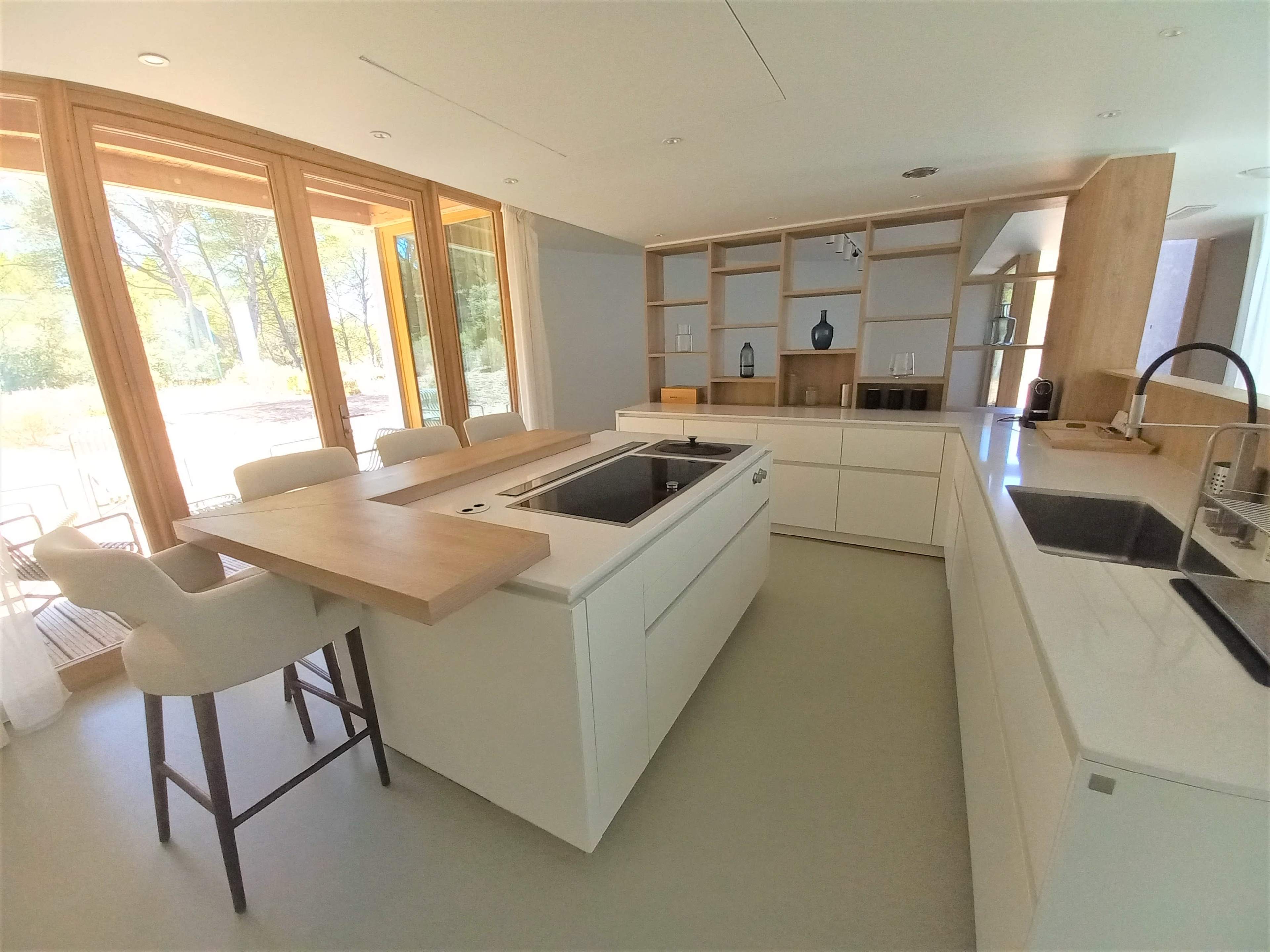 A modern kitchen with an island, white cabinetry, and large windows that overlook a garden.