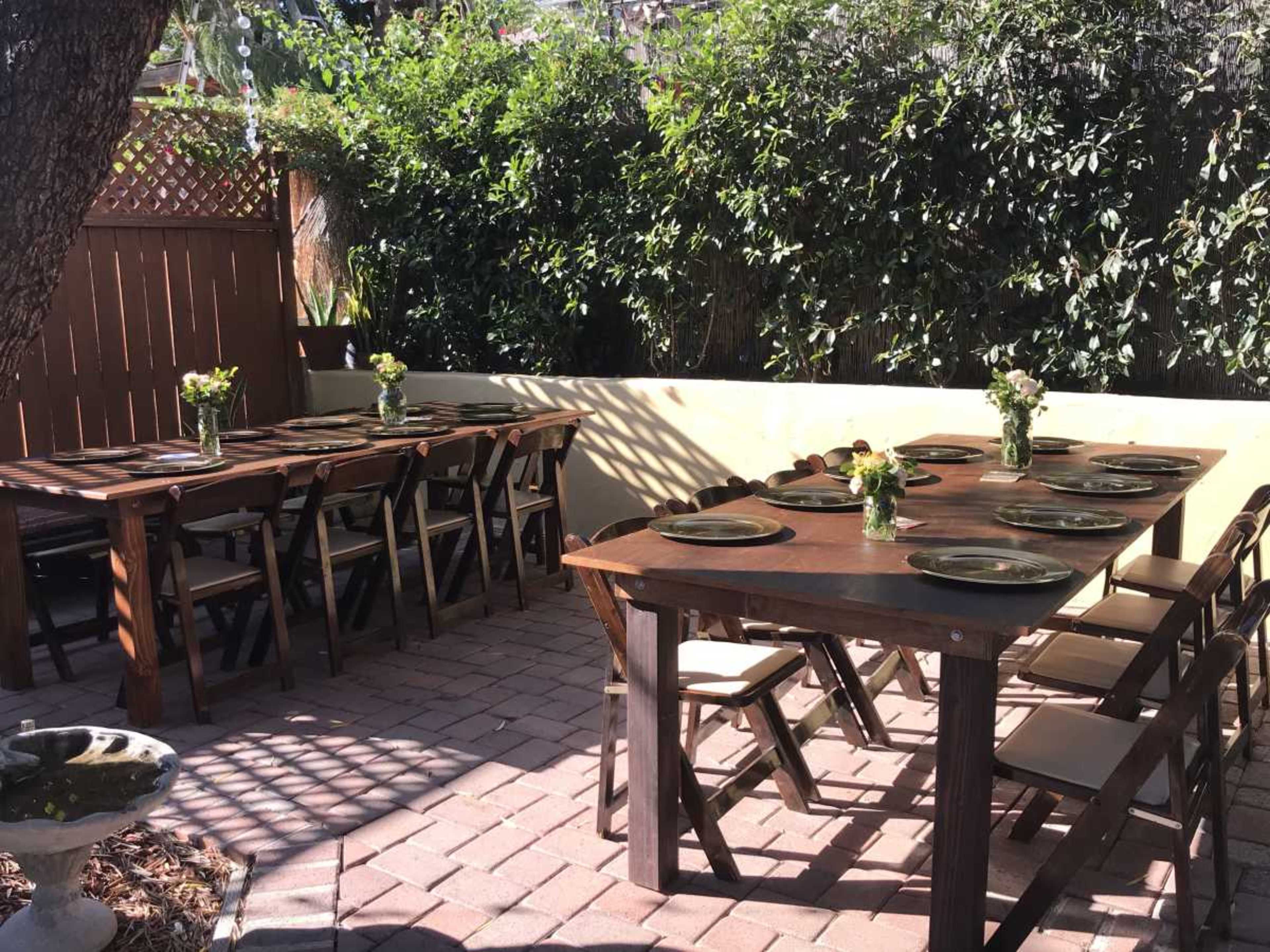 The image shows an outdoor dining area with two wooden tables set with plates and small flower arrangements, surrounded by lush greenery.