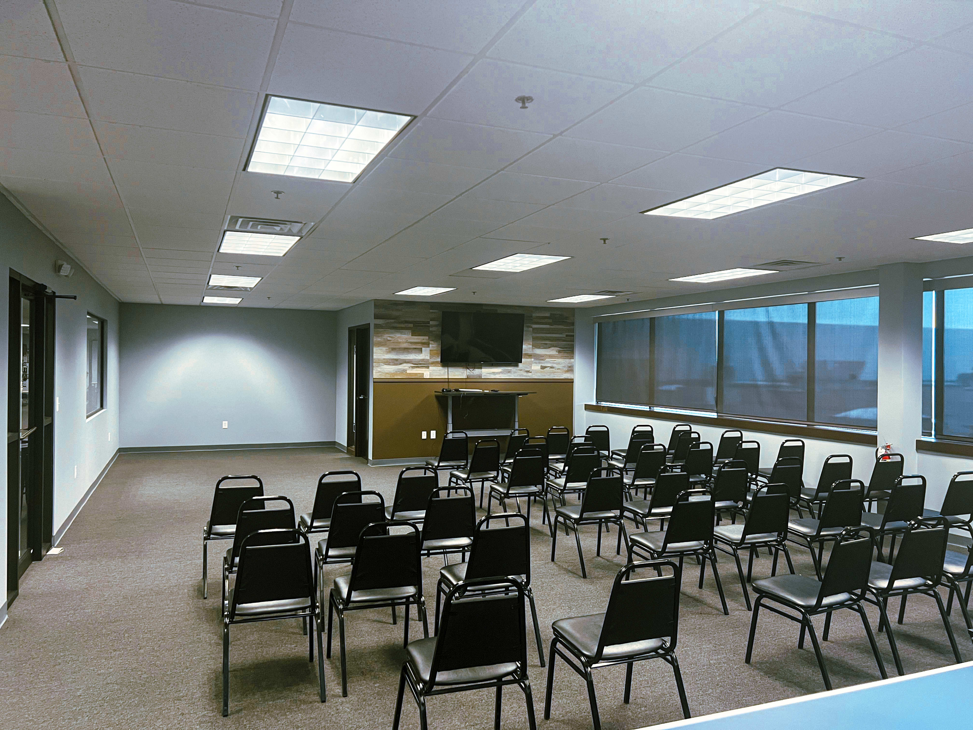 The image shows a conference room set up with rows of black chairs facing a wall-mounted television and a small stage area.