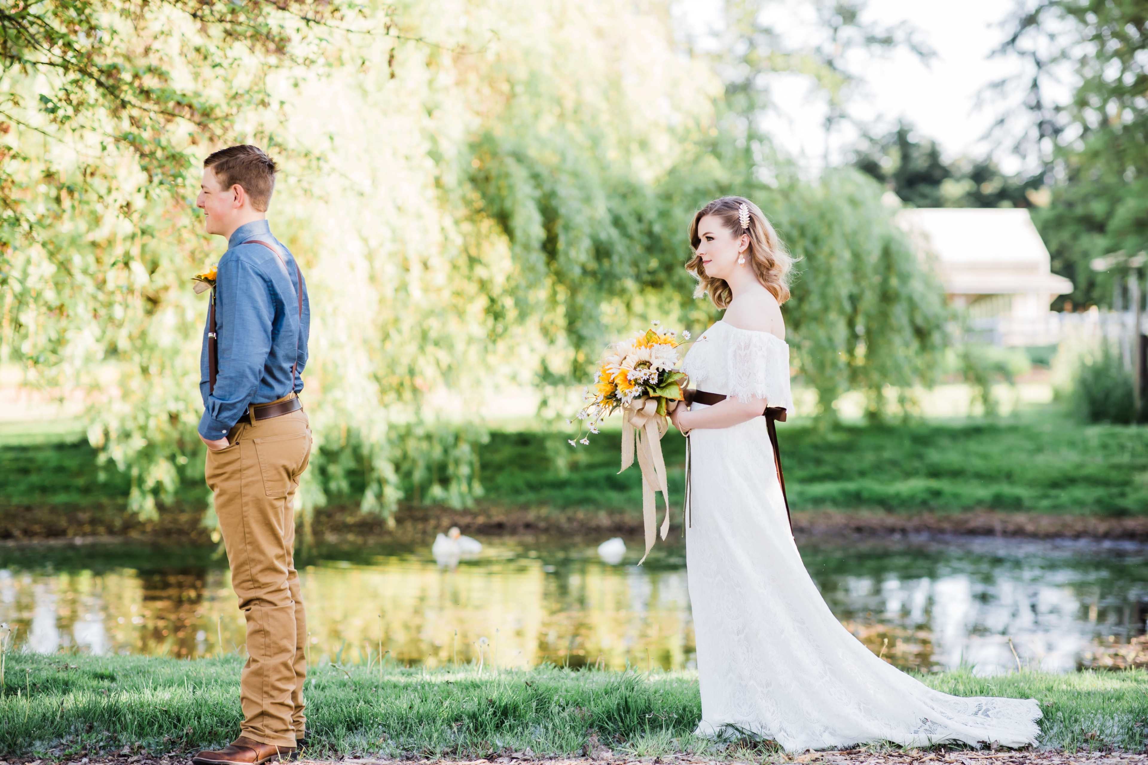 A bride in a white gown holds a bouquet while standing behind a groom, who faces away from her, near a pond with ducks in a sunny outdoor setting.