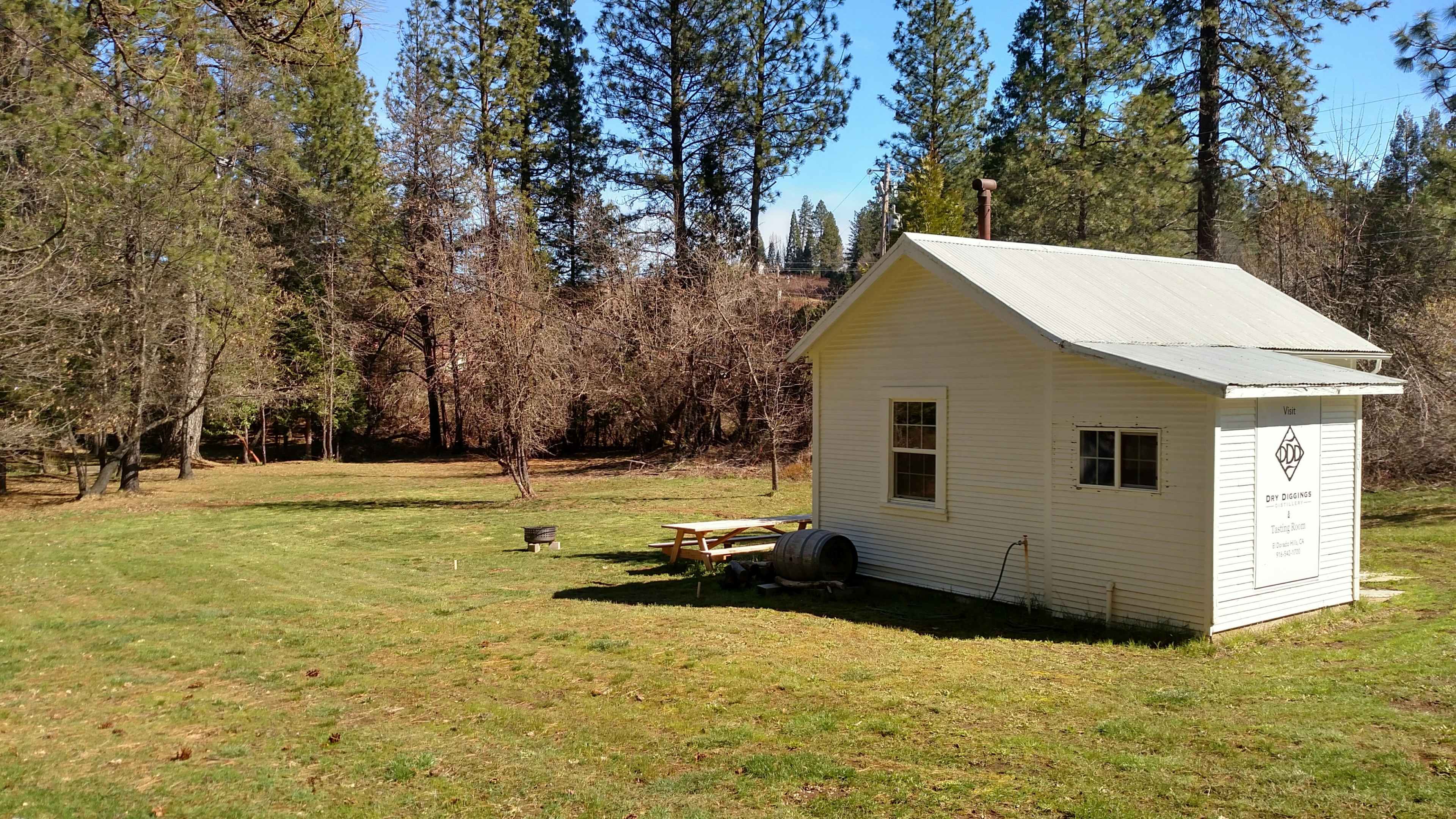 A small white cottage sits on a grassy area surrounded by tall trees and an open field.