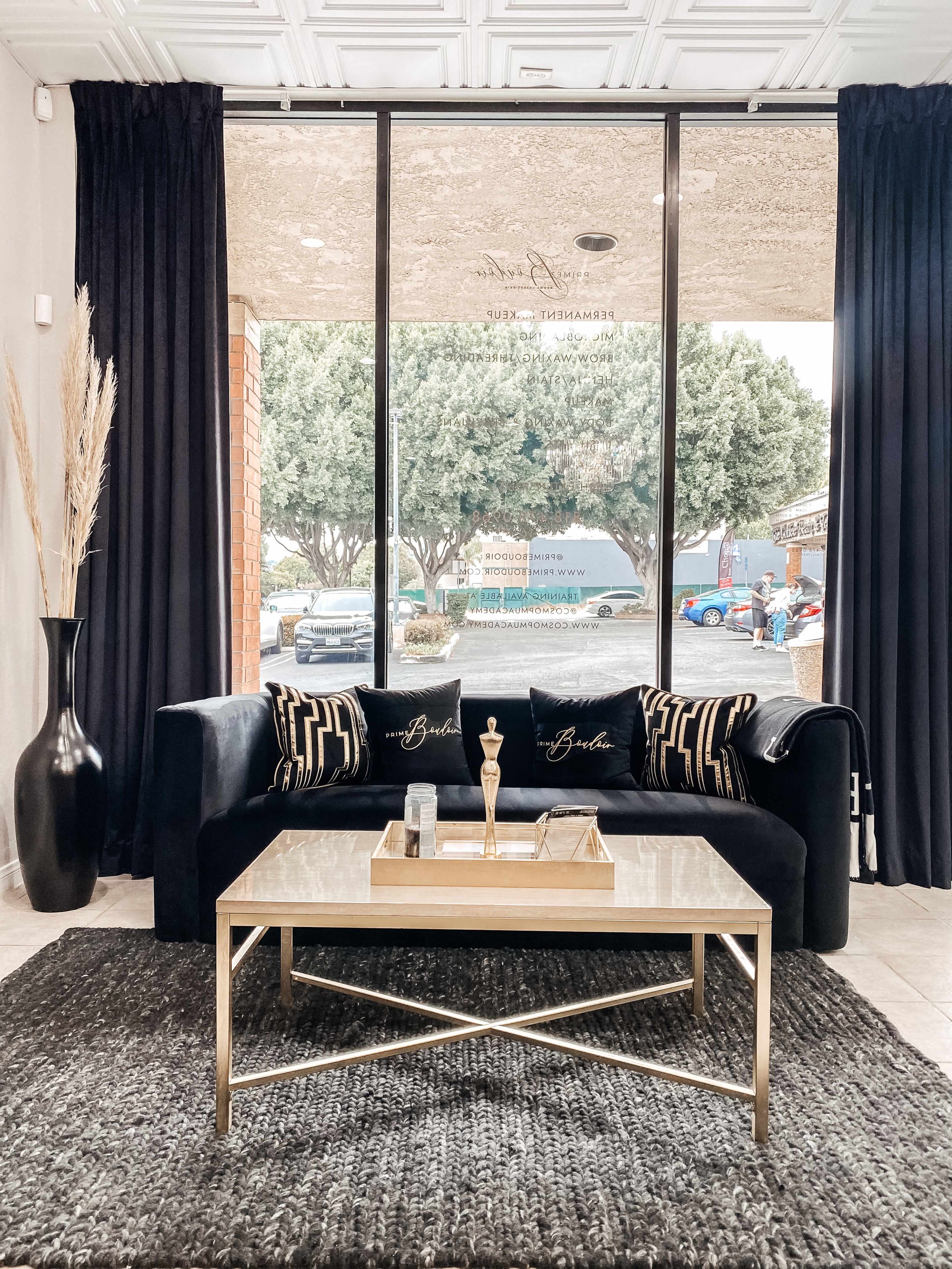 A modern seating area features a black couch with decorative pillows, a glass-top coffee table, and a view of the outdoors through large windows.