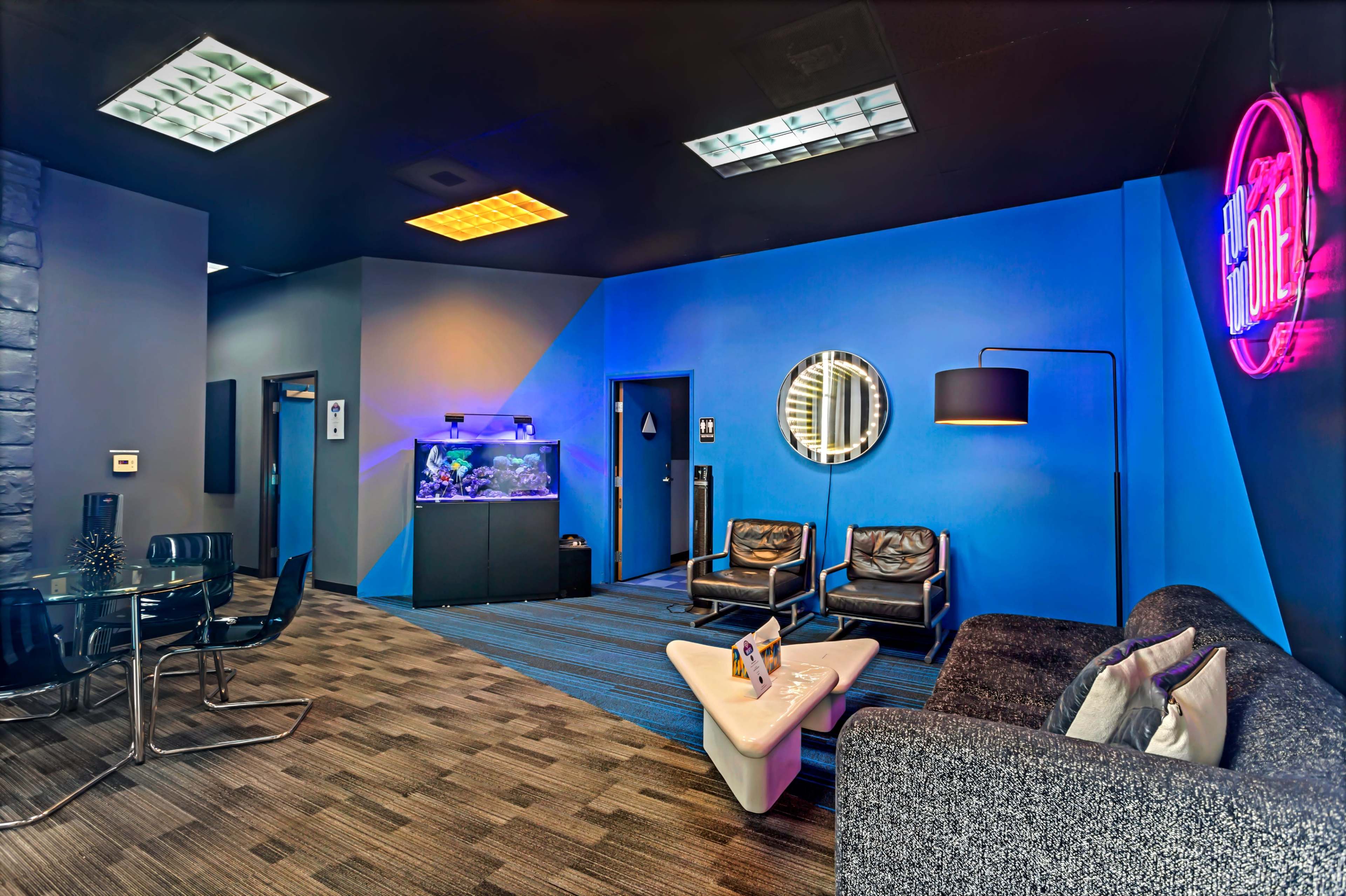 The image shows a modern lounge area with stylish furniture, an aquarium, and blue walls, illuminated by varied lighting.