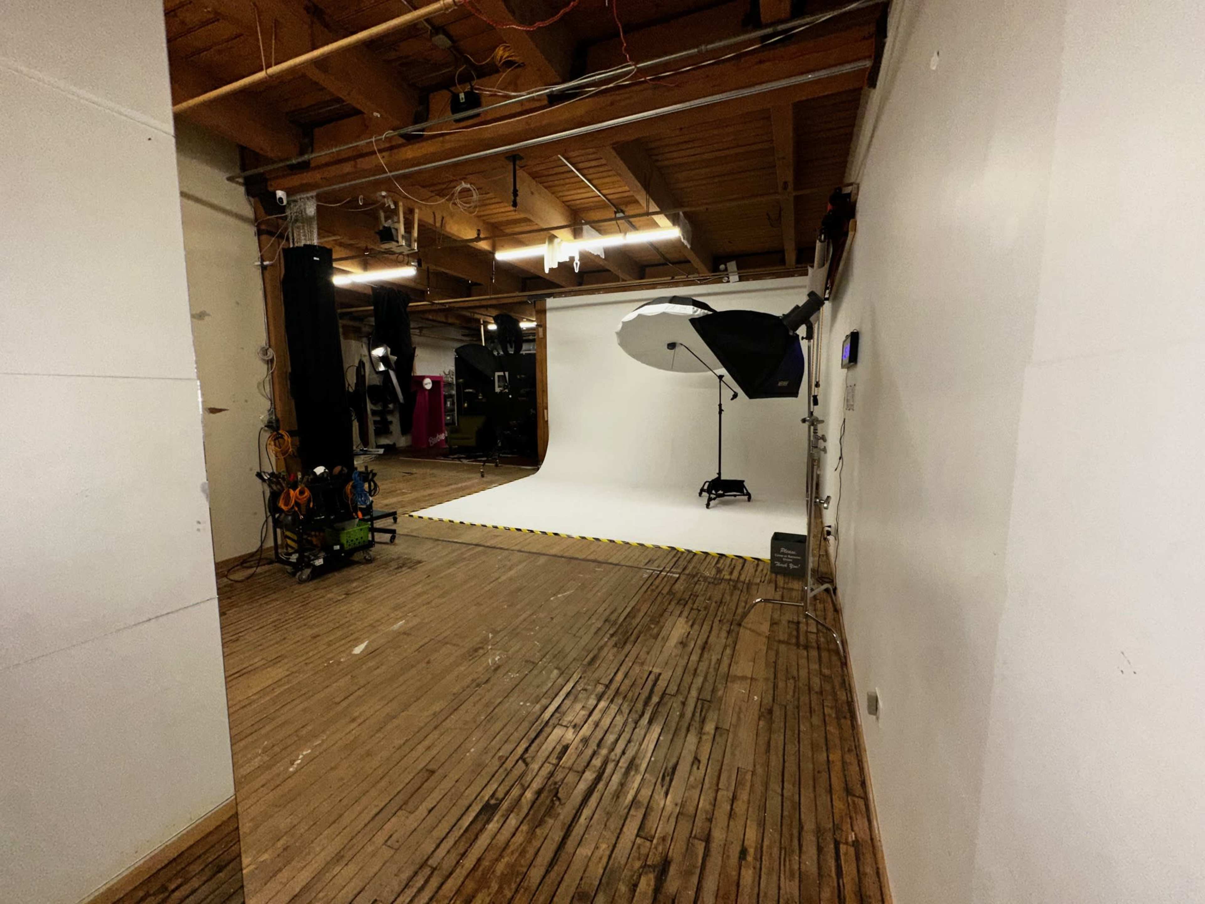 The image shows a photography studio with a white backdrop, wooden flooring, and various equipment, including lights and a camera setup, located in a spacious room.