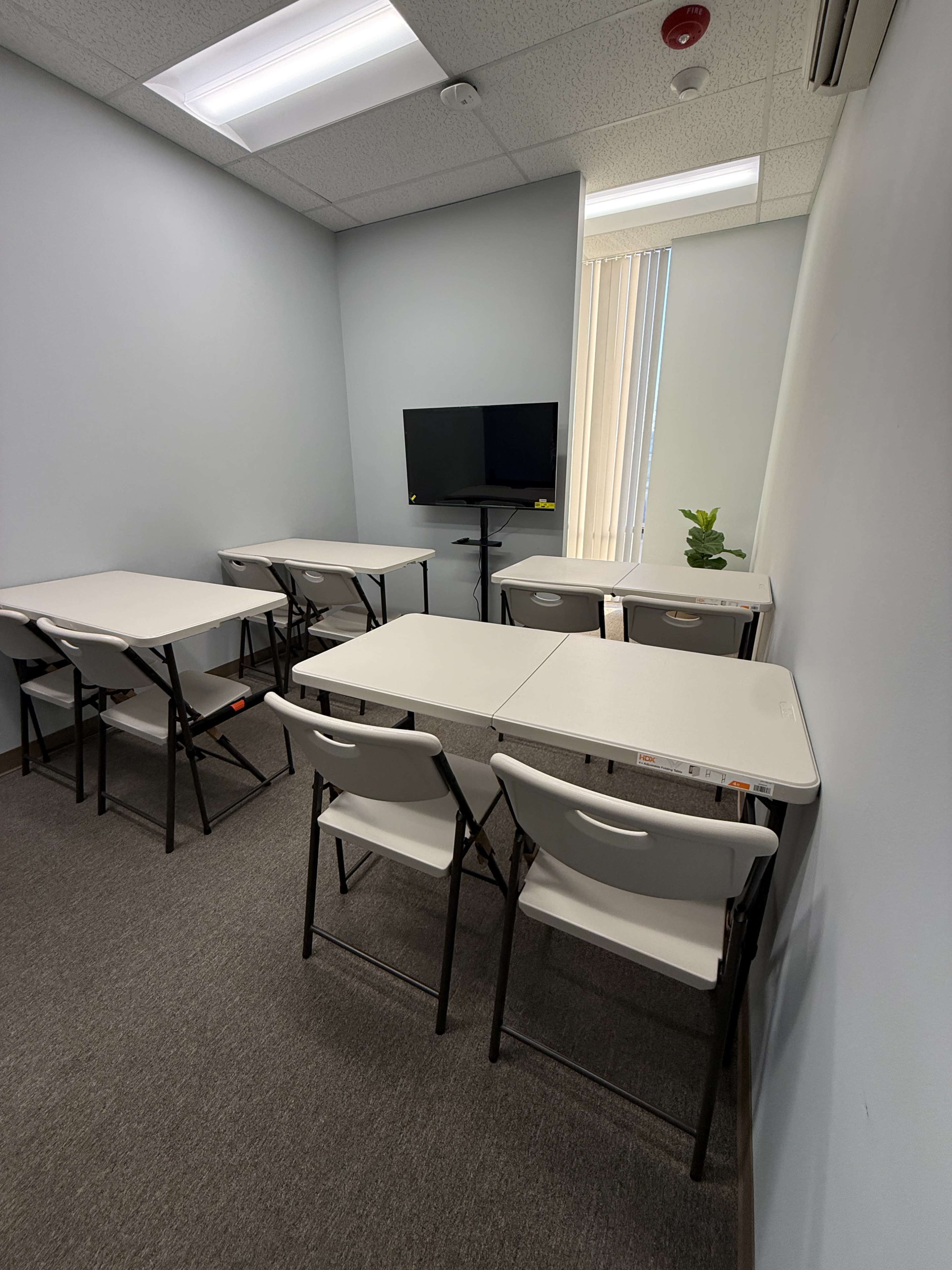 The image shows a small, empty classroom with a television mounted on the wall, several folding tables arranged in a U-shape, and white chairs positioned around the tables.