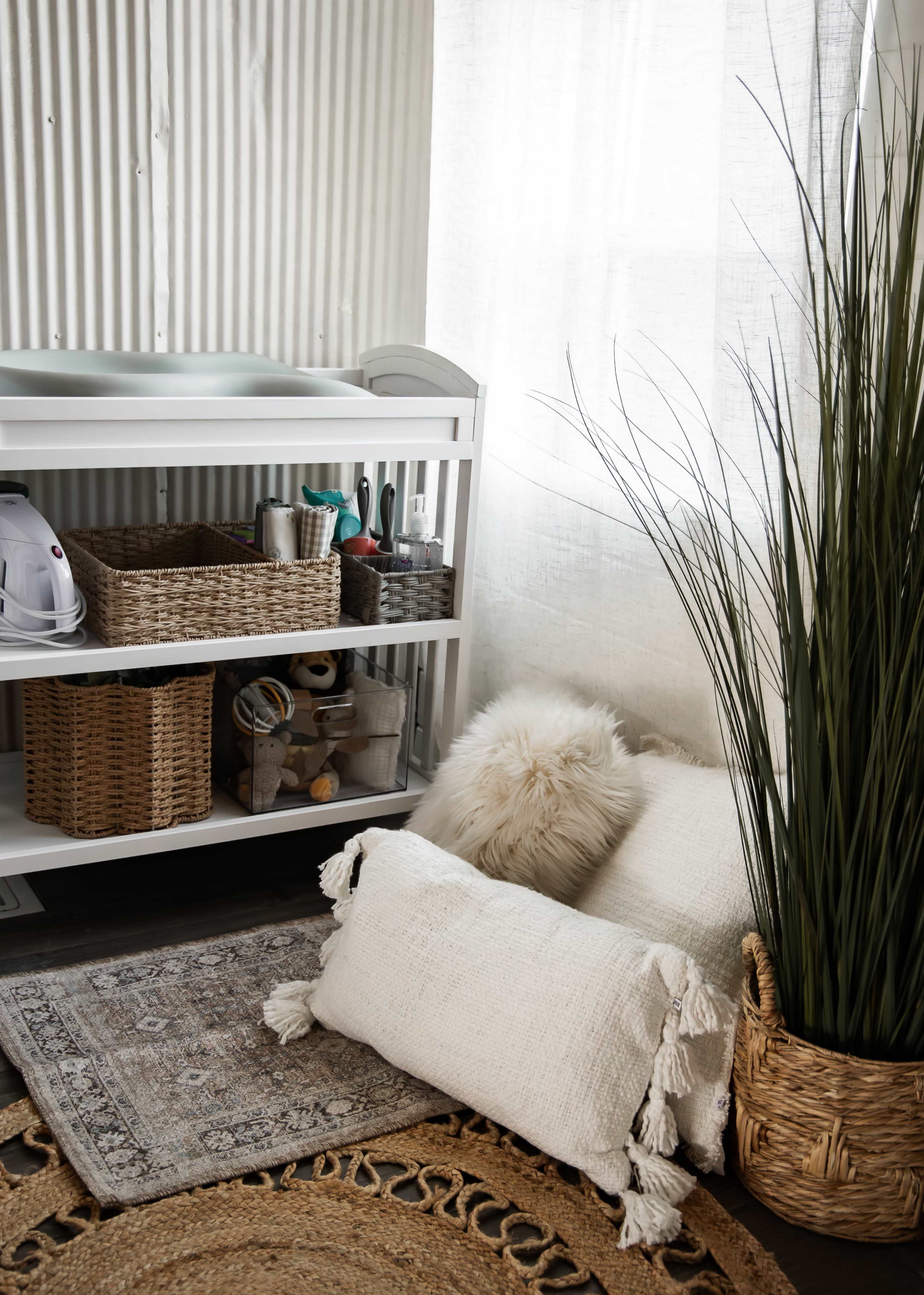 The image shows a styled corner of a room with a changing table, baskets, plush pillows, and decorative plants.