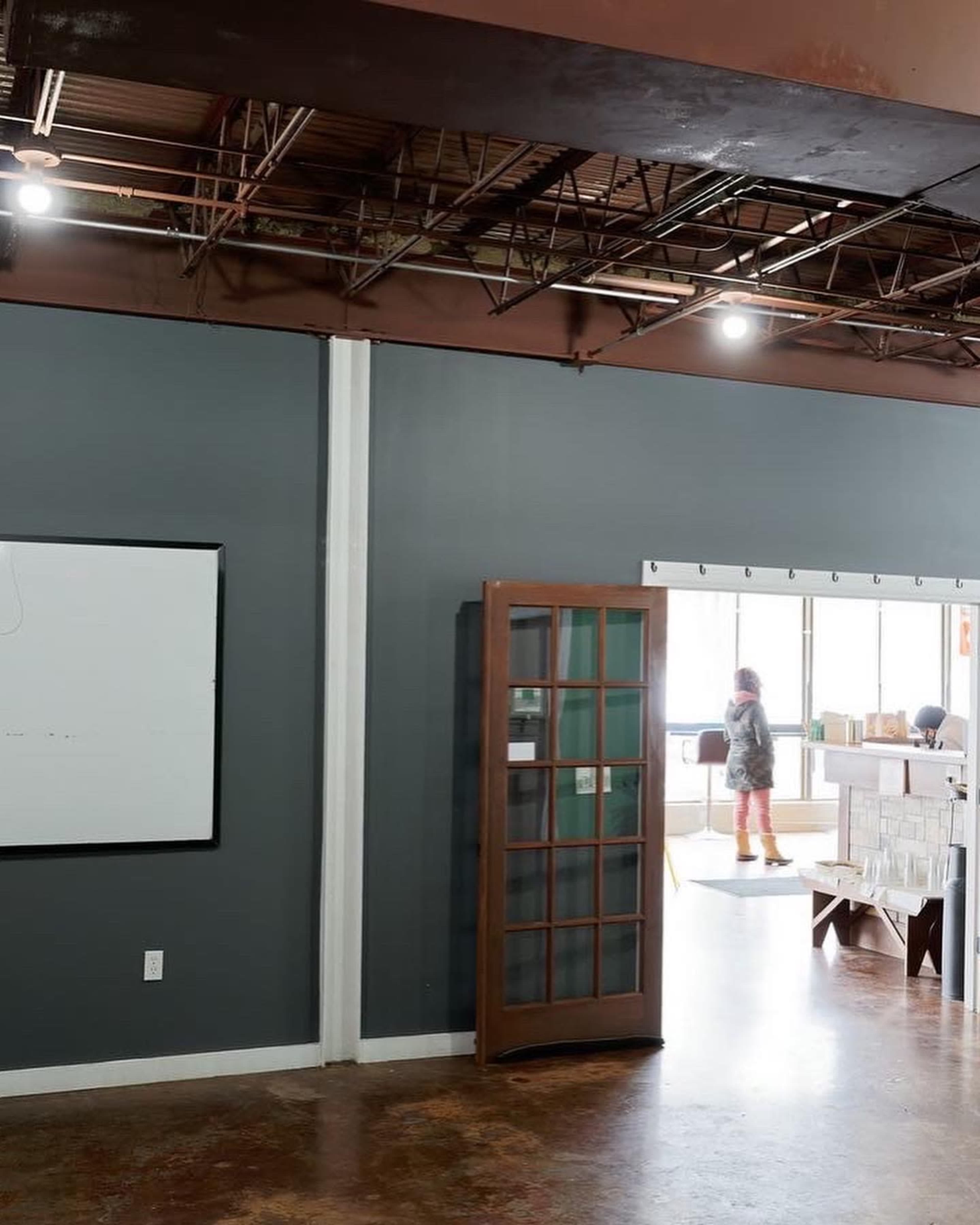 The image shows an open office space with a whiteboard on the wall and a wooden door leading to another area where a person is standing.