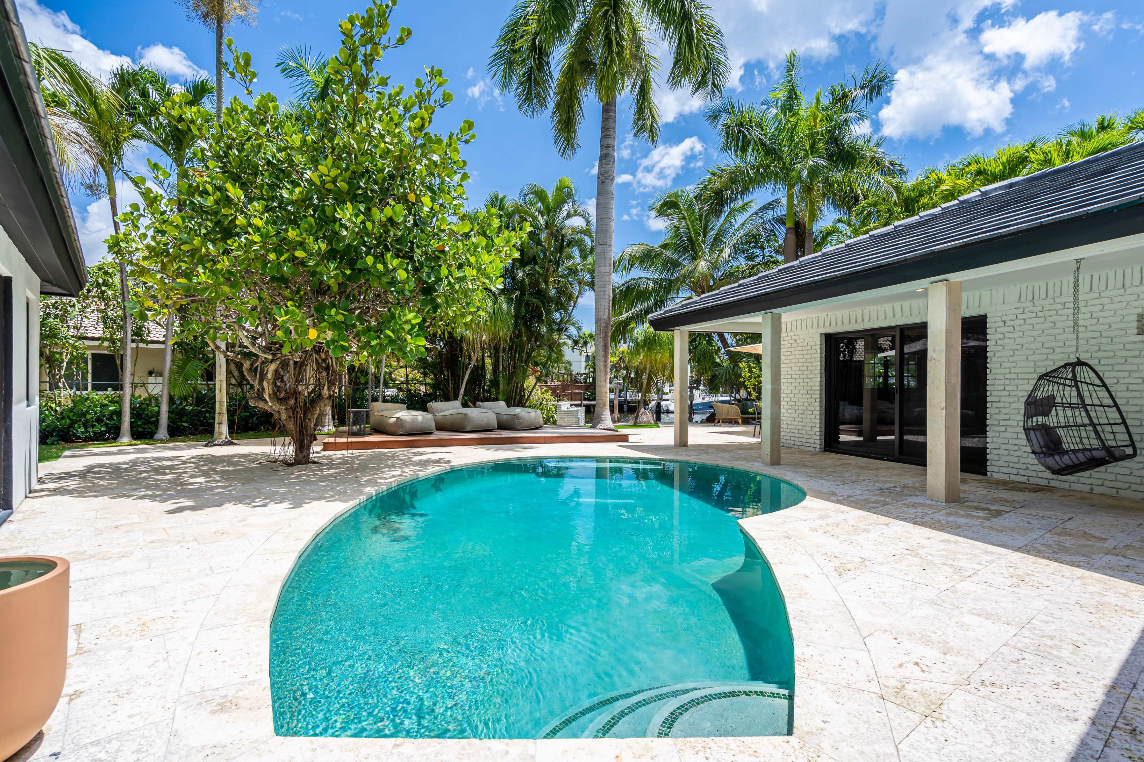 A curved swimming pool surrounded by palm trees and a patio area with a hanging chair and outdoor seating.