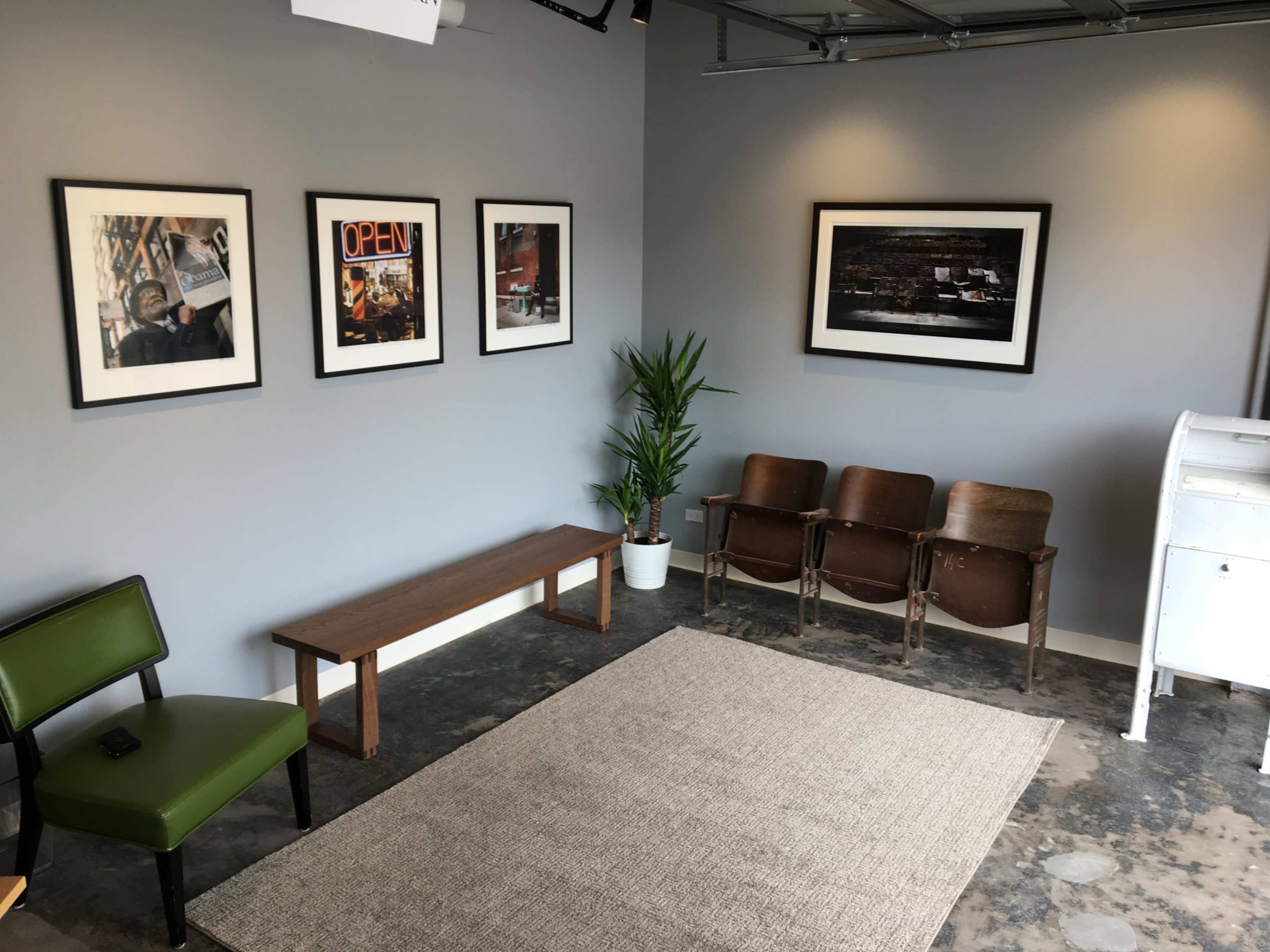 The image shows a small waiting area with a green chair, a wooden bench, and two brown chairs, decorated with framed photographs on the wall and a potted plant in the corner.