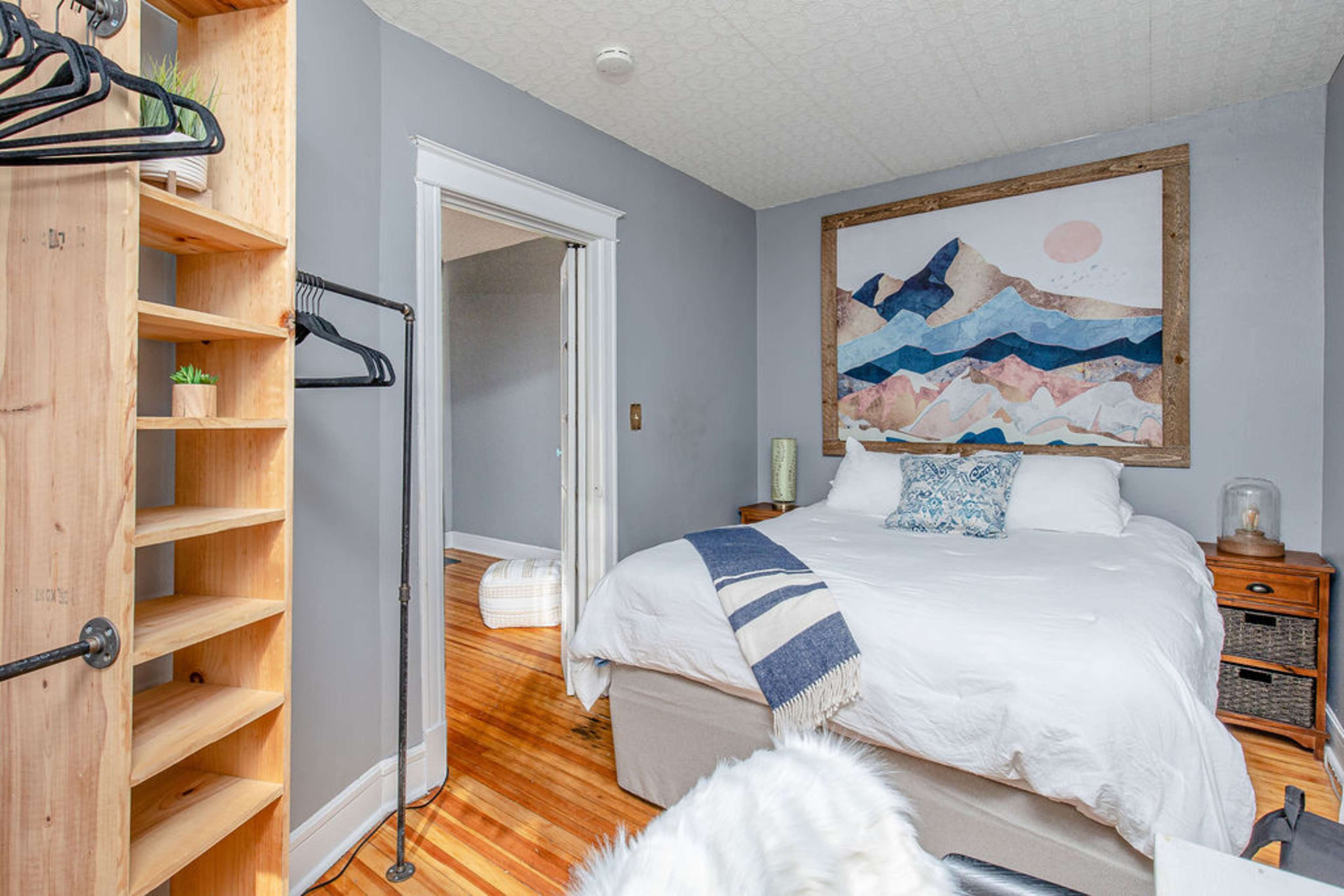 A small bedroom features a bed with white linens, a large framed artwork above it, a wooden shelf, and a doorway leading to another room.