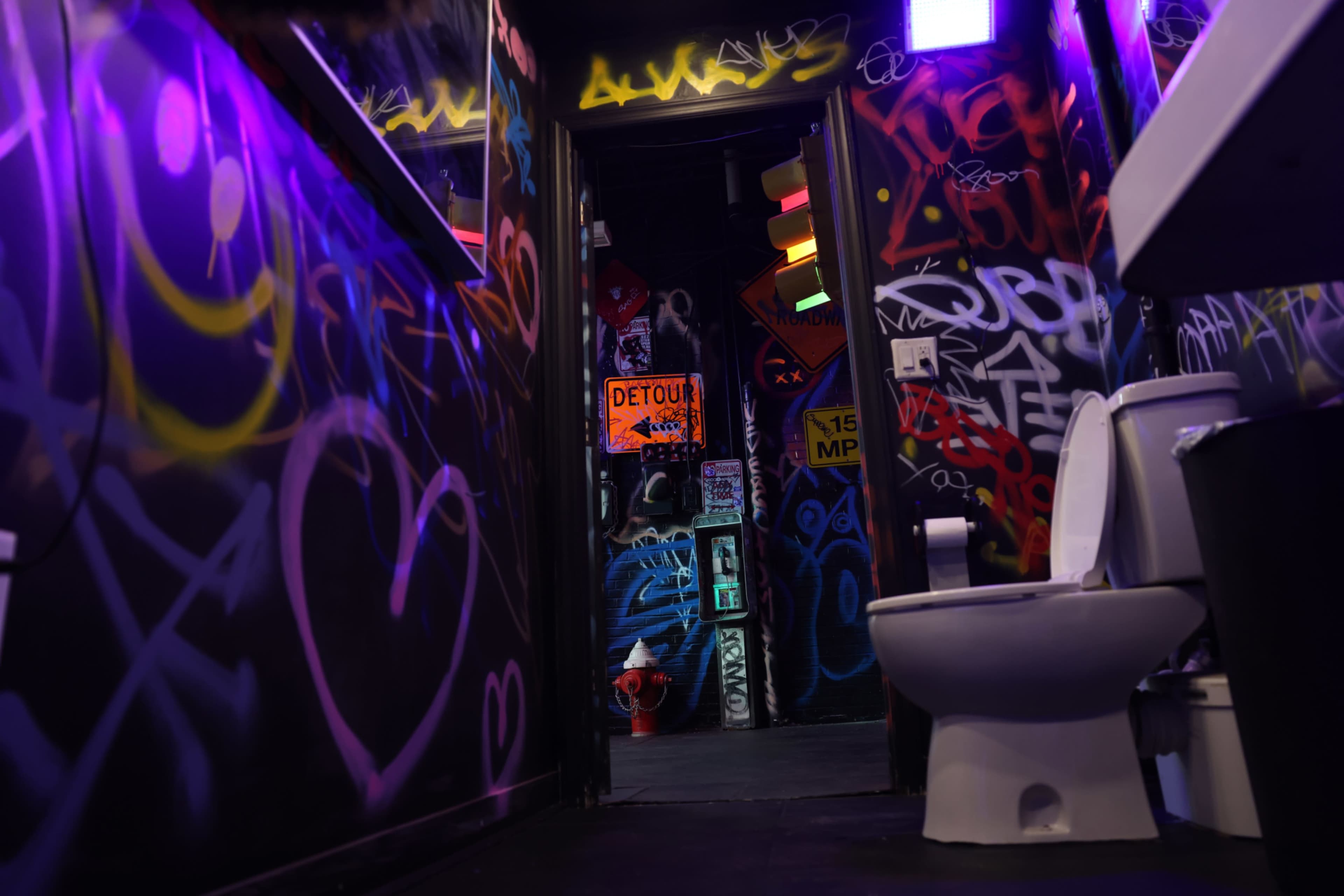 The image captures a dimly lit bathroom entrance characterized by colorful graffiti on the walls and a white toilet positioned prominently in the foreground.