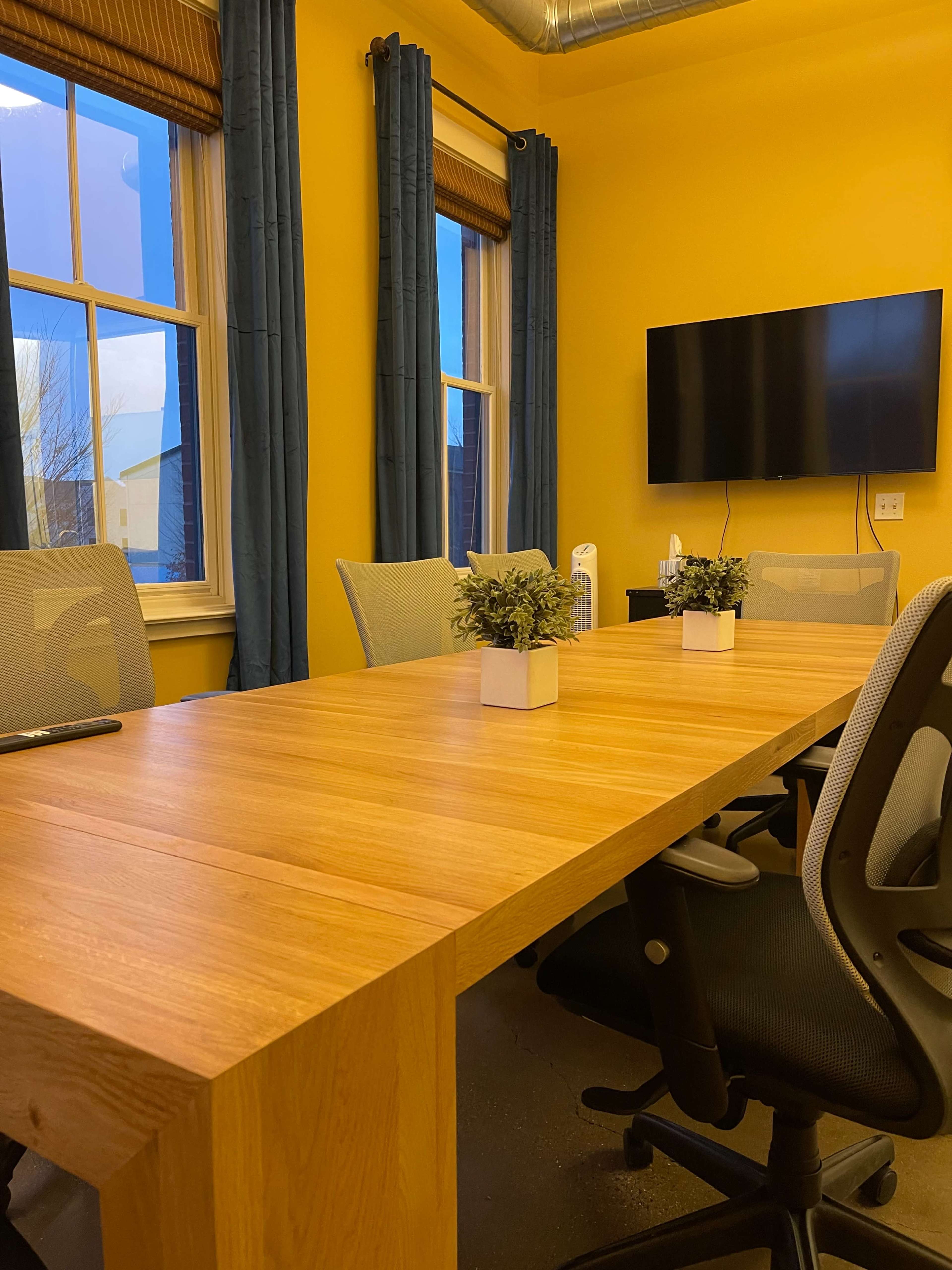 The image shows a conference room with a long wooden table, four chairs, and potted plants centered along the table, with yellow walls and a large TV mounted on the wall.