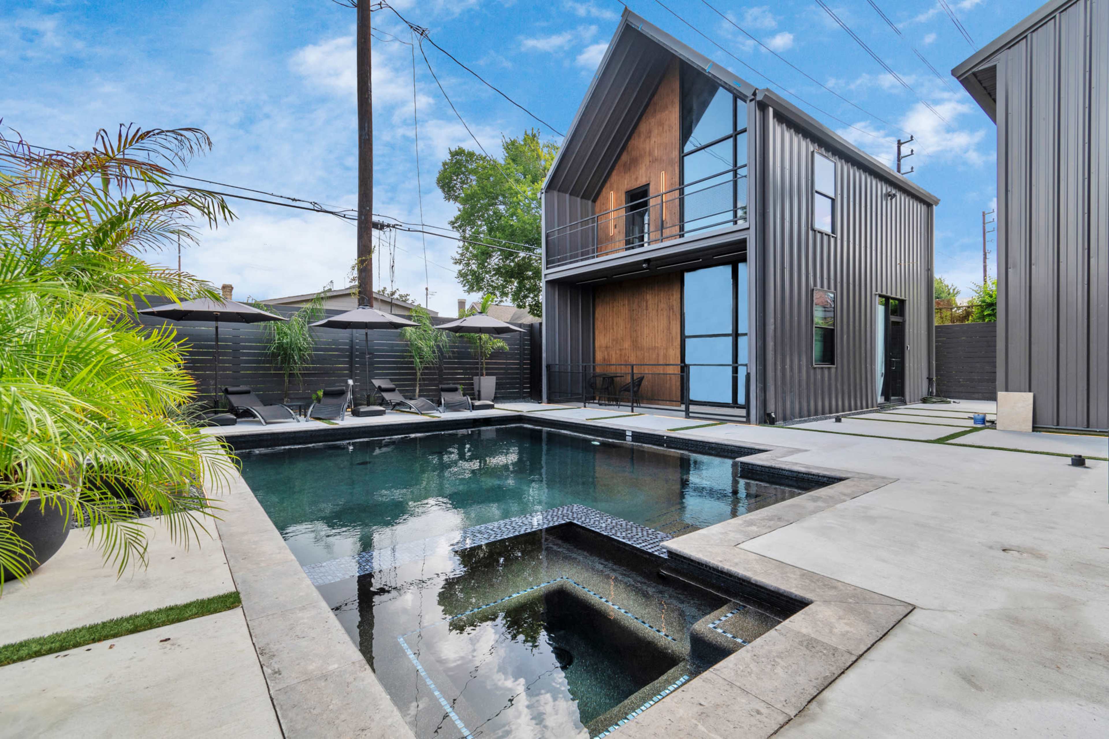 A modern outdoor space featuring a rectangular swimming pool surrounded by concrete and landscaping, with two contemporary buildings in the background.