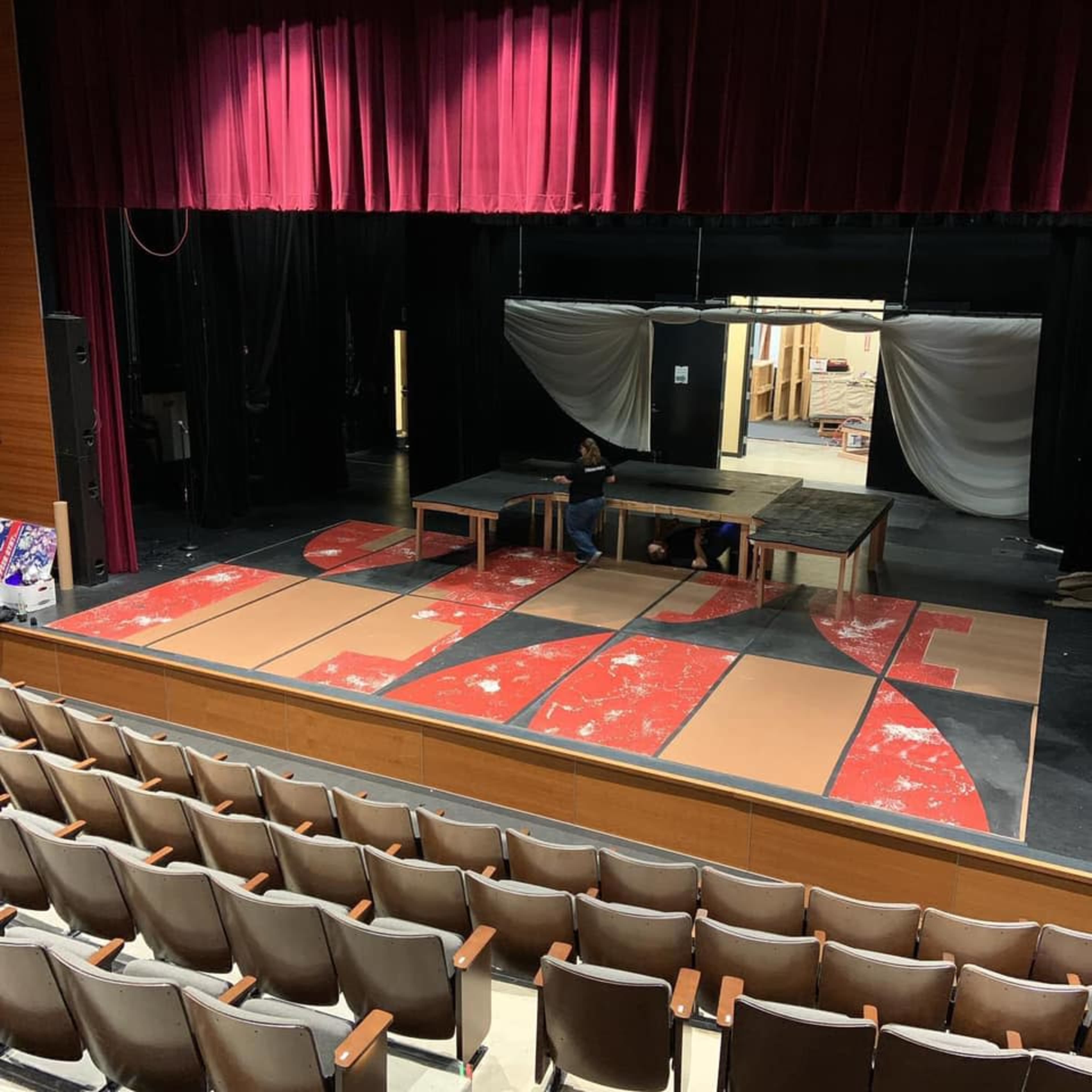 A stage is set up with wooden tables and large painted panels on the floor, while people work in the background preparing for a performance.