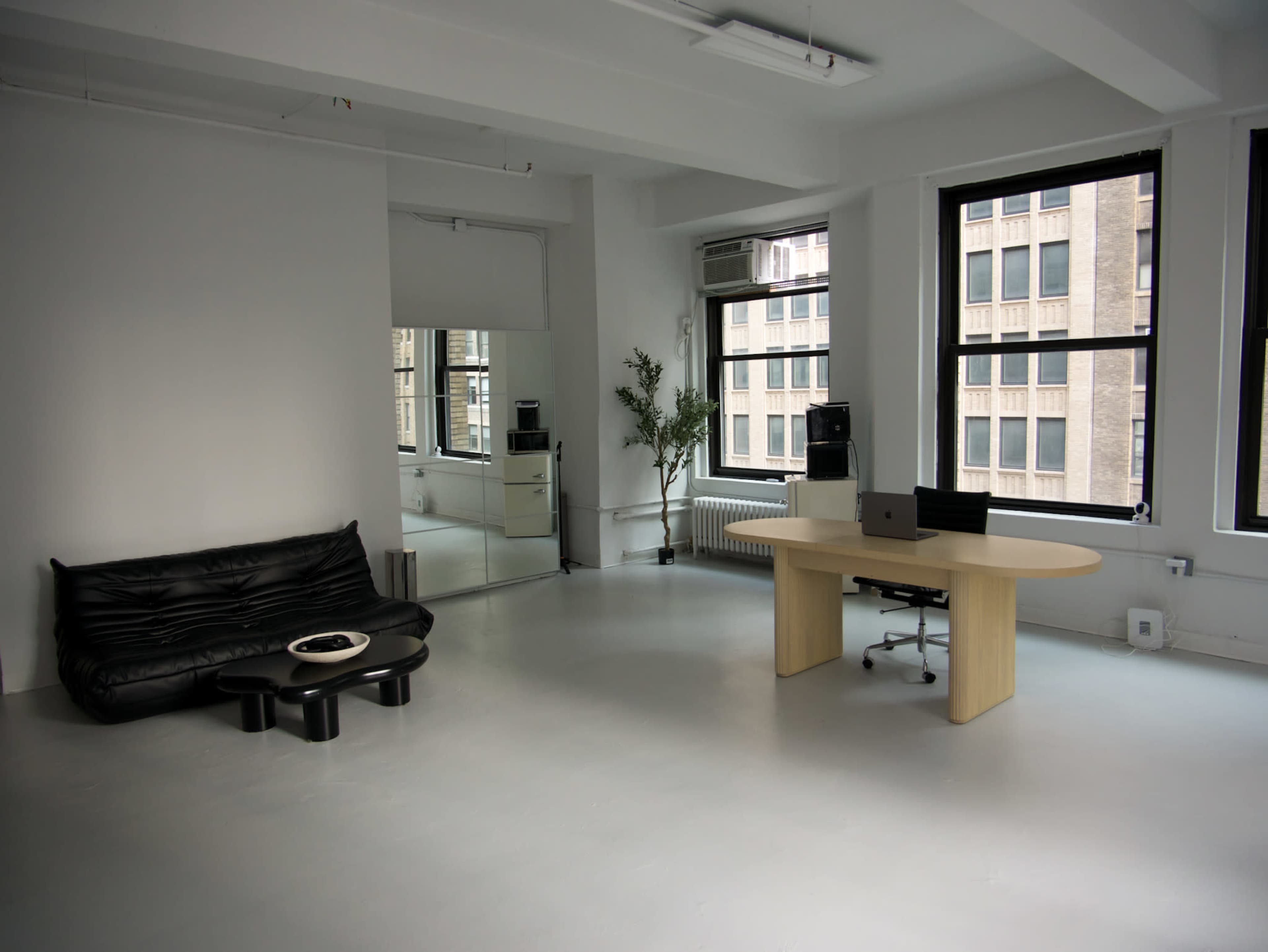The image shows a minimalist office space with a black sofa, a round coffee table, and a wooden desk with a chair near large windows.