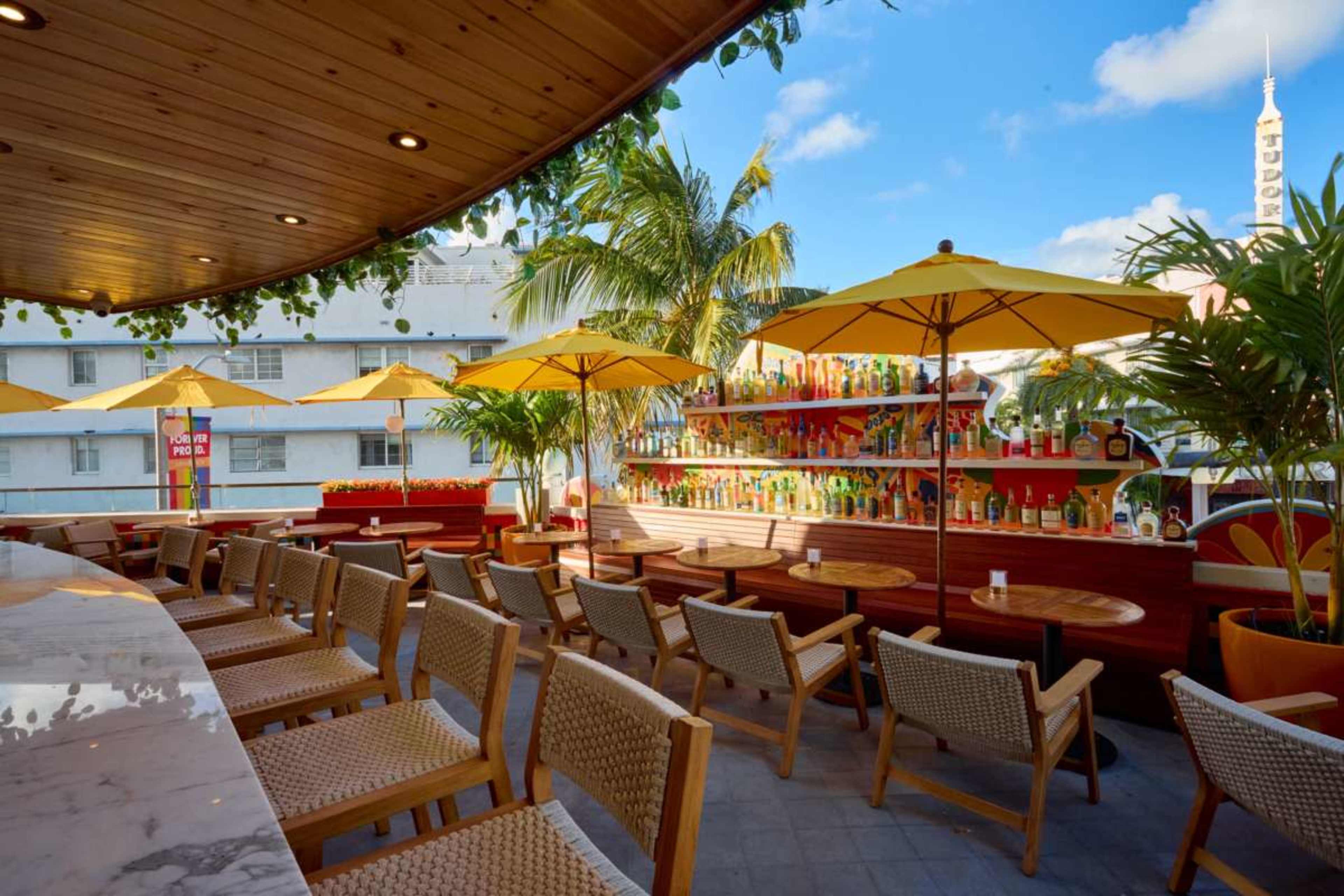 The image depicts an outdoor bar area with wooden seating, yellow umbrellas, and a colorful display of bottles on shelves in the background.